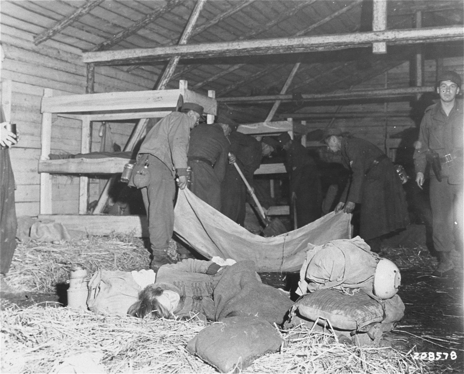 Medical corpsmen of the U.S. 71st Infantry Division, 3rd U.S. Army look on as captured German soldiers remove bodies from inside a barracks in Gunskirchen.  In the foreground, a Jewish girl lies huddled in the straw on the floor of the barracks.