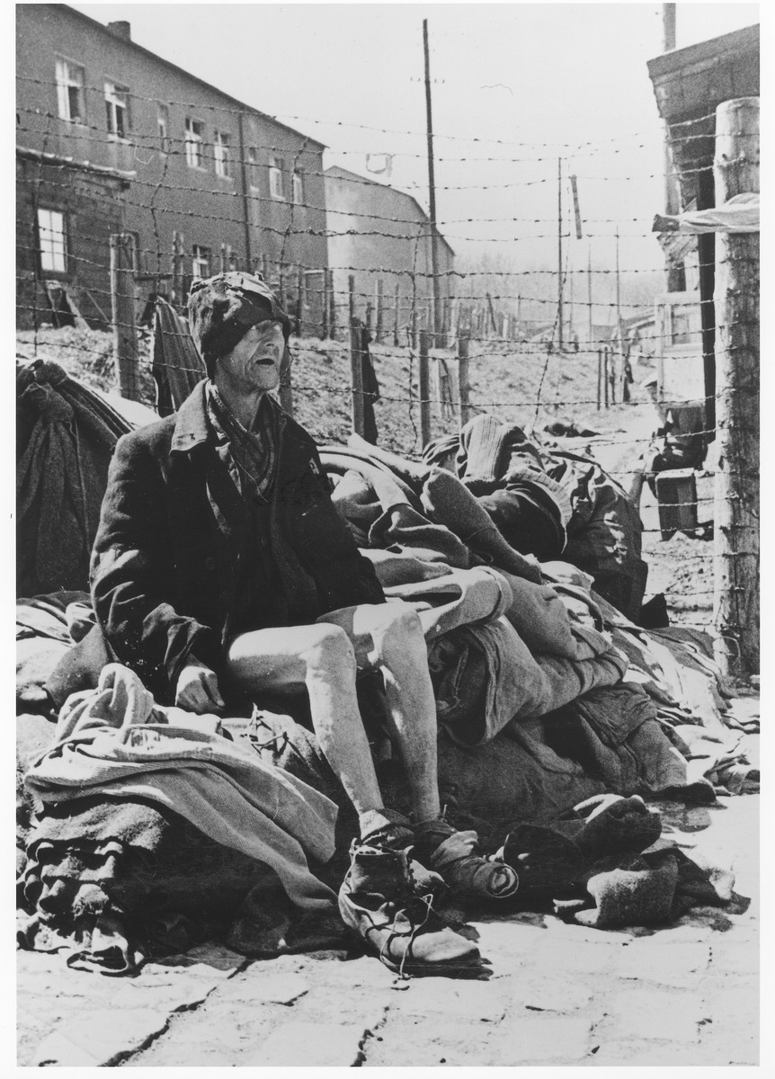 Close-up of an emaciated middle-aged survivor, wearing a tattered cap, sits on a pile of blankets alongside a barbed wire fence in Buchenwald.  He had been imprisoned in Buchenwald for seven years.  Behind him, another survivor rests on the same pile.
