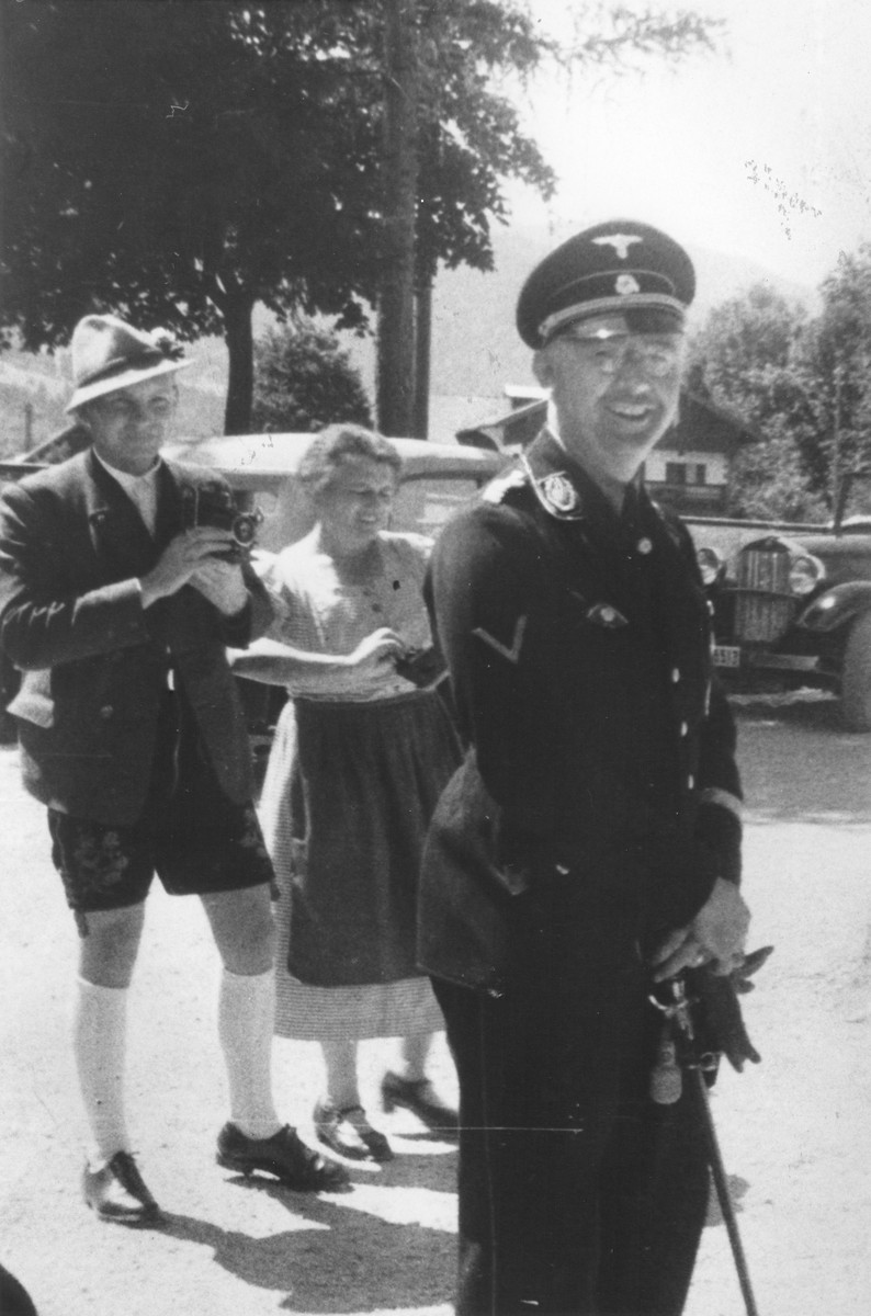 Reichsfuehrer-SS Heinrich Himmler stands outside in front of a couple with a camera.