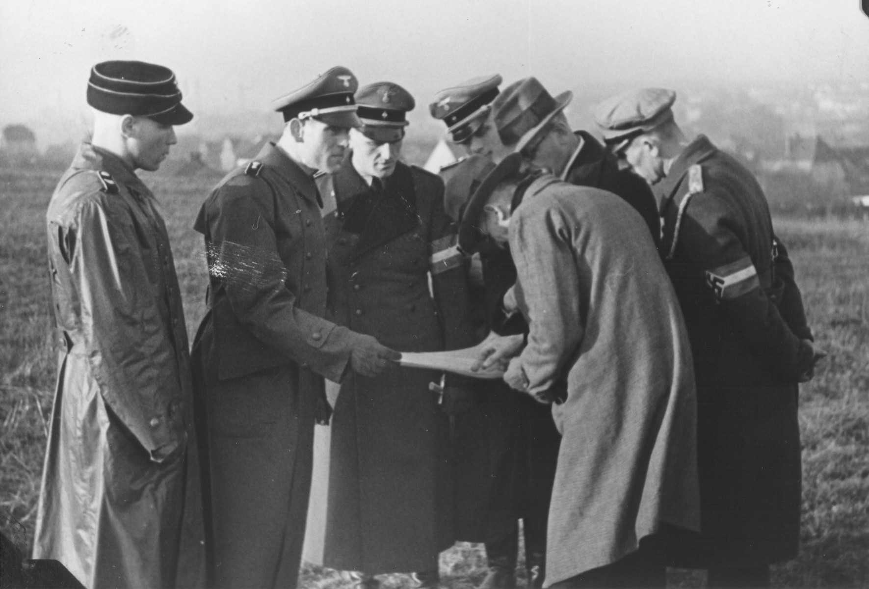 A group of uniformed Germans look on as two civilians study a map in an open field.