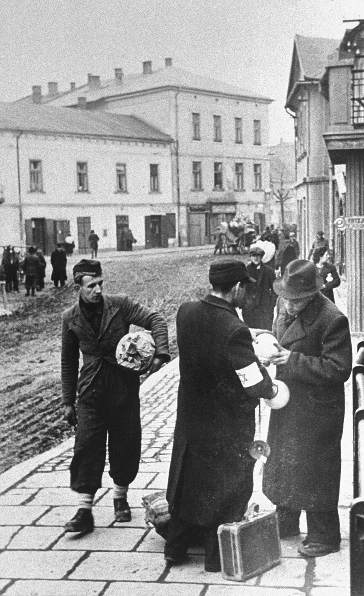 Two Jewish men examine lighting fixtures on the street in the Krakow ghetto, while a third man walks by holding a bundle under his arm.