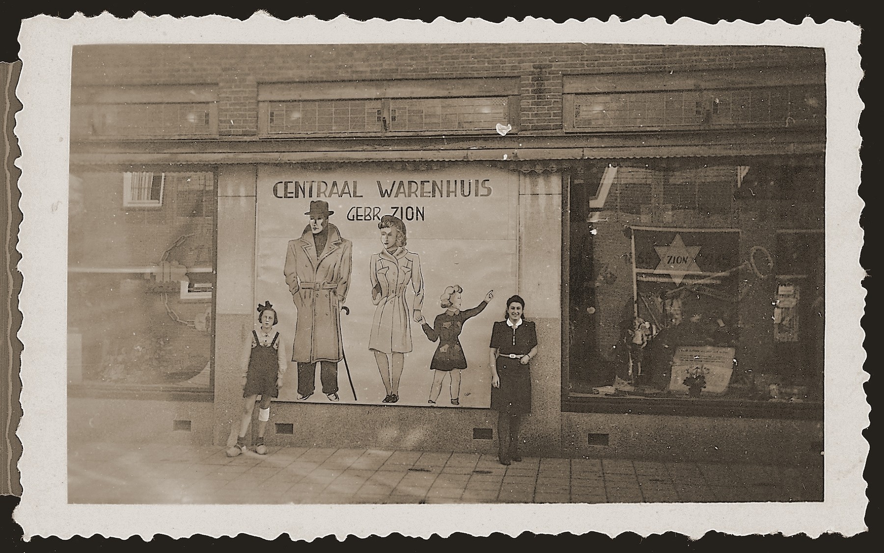 Bep Meijer (right) poses next to a billboard in the window of the newly reopened Zion clothing and fabric store in Eibergen.  The sign reads "Central Warehouse, Zion Brothers."