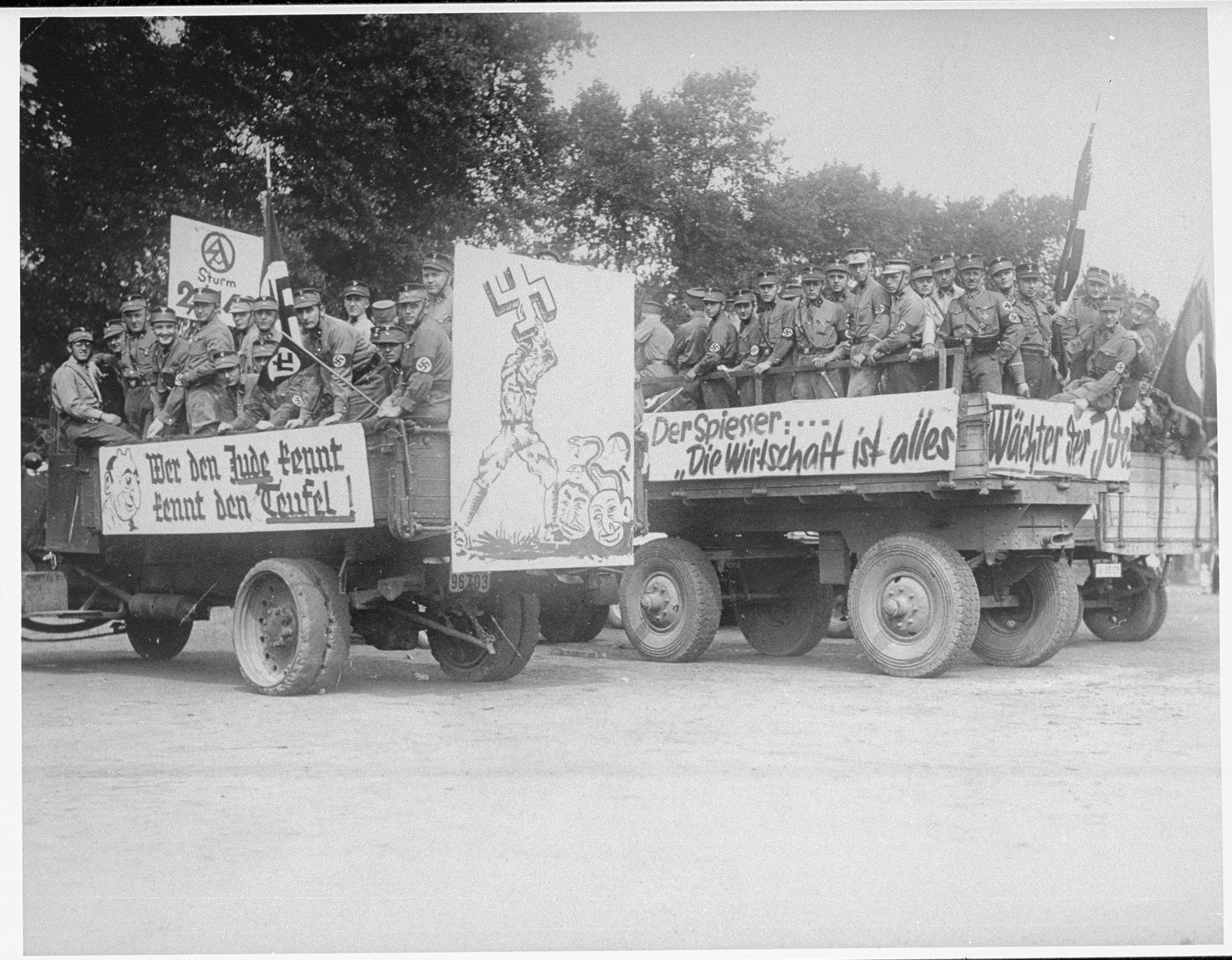 Members of the SA drive through the streets of Recklingshausen, Germany on propaganda trucks bedecked with anti-Jewish banners.

The banners read: "He who knows the Jew knows the Devil,"  "The Bourgeois: 'the economy is everything."  The poster depicts a man using a swastika to crush Jews with snakes coming out of their heads.  On the back is the official stamp of the SA and a handwritten note, "At the beginning of the Propaganda parade on August 18, 1935."