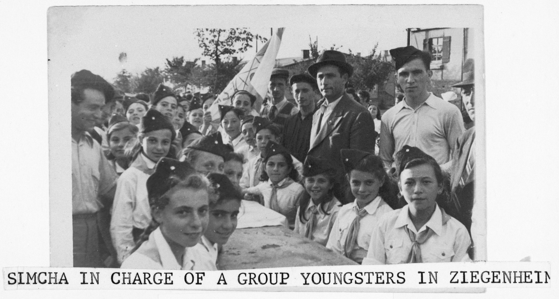 Children in the Ziegenhain displaced persons camp sit outside around a table in their new youth movement uniforms.

Posing with them are their two instructors, Simcha Langzam and Abraham Lomp.