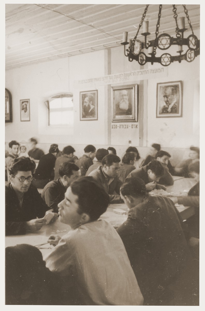 Members of Kibbutz Nili hachshara (Zionist collective) in Pleikershof, Germany, sit around tables in the dining hall.

On the wall are photographs of Herzl, Weizman and A.D. Gordon and the Hebrew motto, "Man-Work-Nature."