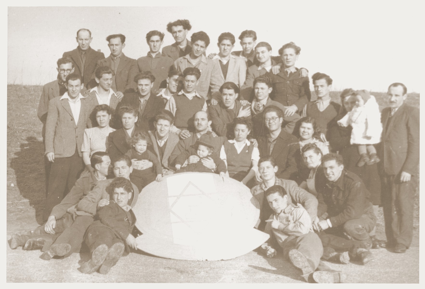 Group portrait of the members of the Kibbutz Nili hachshara (Zionist collective) in Pleikershof, Germany, posing around a large plaque inscribed with a Star of David and the Hebrew word "Nachem" (comfort).  

Seated in the center is Noach Miedzinski holding his daughter Nili.  On the far right is Aaron Charny standing next to his daughter.