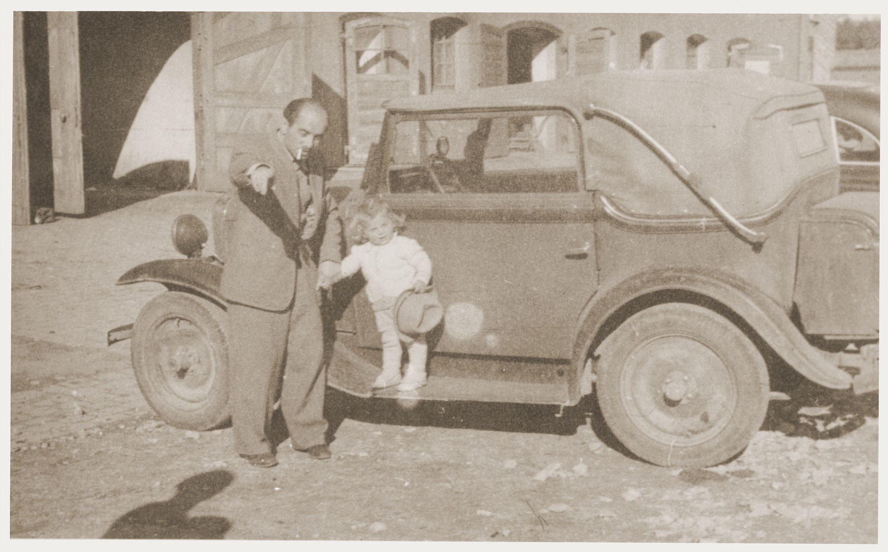 A little girl poses on the running board of a car while holding her father's hand at the Kibbutz Nili hachshara (Zionist collective) in Pleikershof, Germany.  

Pictured are Noach Miedzinski and his daughter Nili Ruchana.