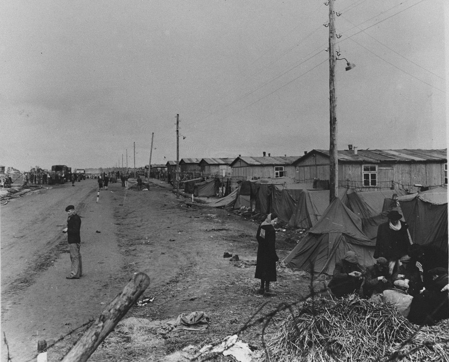 View of the main street in the Bergen-Belsen concentration camp (looking toward the entrance) lined with a row of small tents that have been pitched outside the barracks.  

In the foreground, a group of survivors huddles in front of one of the tents, while, in the distance, other liberated prisoners walk along the unpaved road. 

Original caption is illegible.