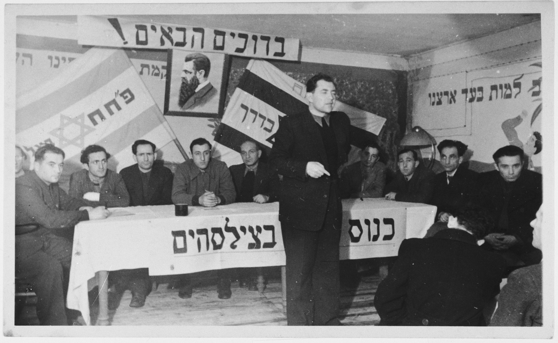 Zionist meeting of the Partizanim-Hayyalim-Halutzim in the Zeilsheim displaced persons' camp held in a room bedecked with a flag, posters and a portrait of Theodore Herzl.