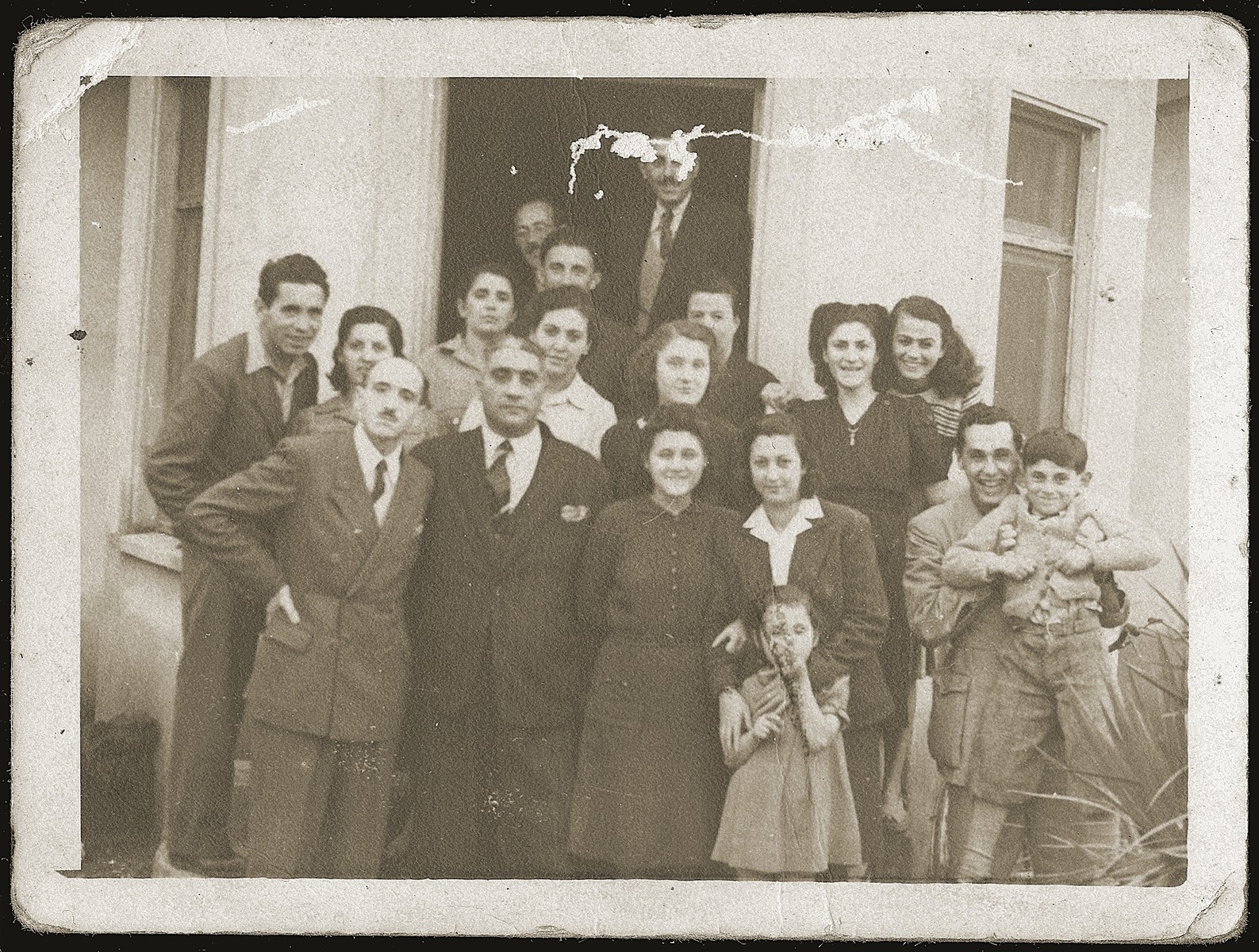 Group portrait of Jewish refugees in front of the home of Neshed Ismail in Tirana after the liberation.  

Pictured are Mosa (Moshe) Mandil (top, in the doorway), Gabriela Mandil (center, middle row),Irena (front) and Gavra Mandil (at the far right) and Neshed Ismail (far left).  Also pictured are Mr. and Mrs Ruchvarger; Mr. and Mrs. Sabitai Gershon and their daughter Stella; Yosef, Finitza and Rozica Ben-Yosif; and Mr. and Mrs. Naftali Komforti