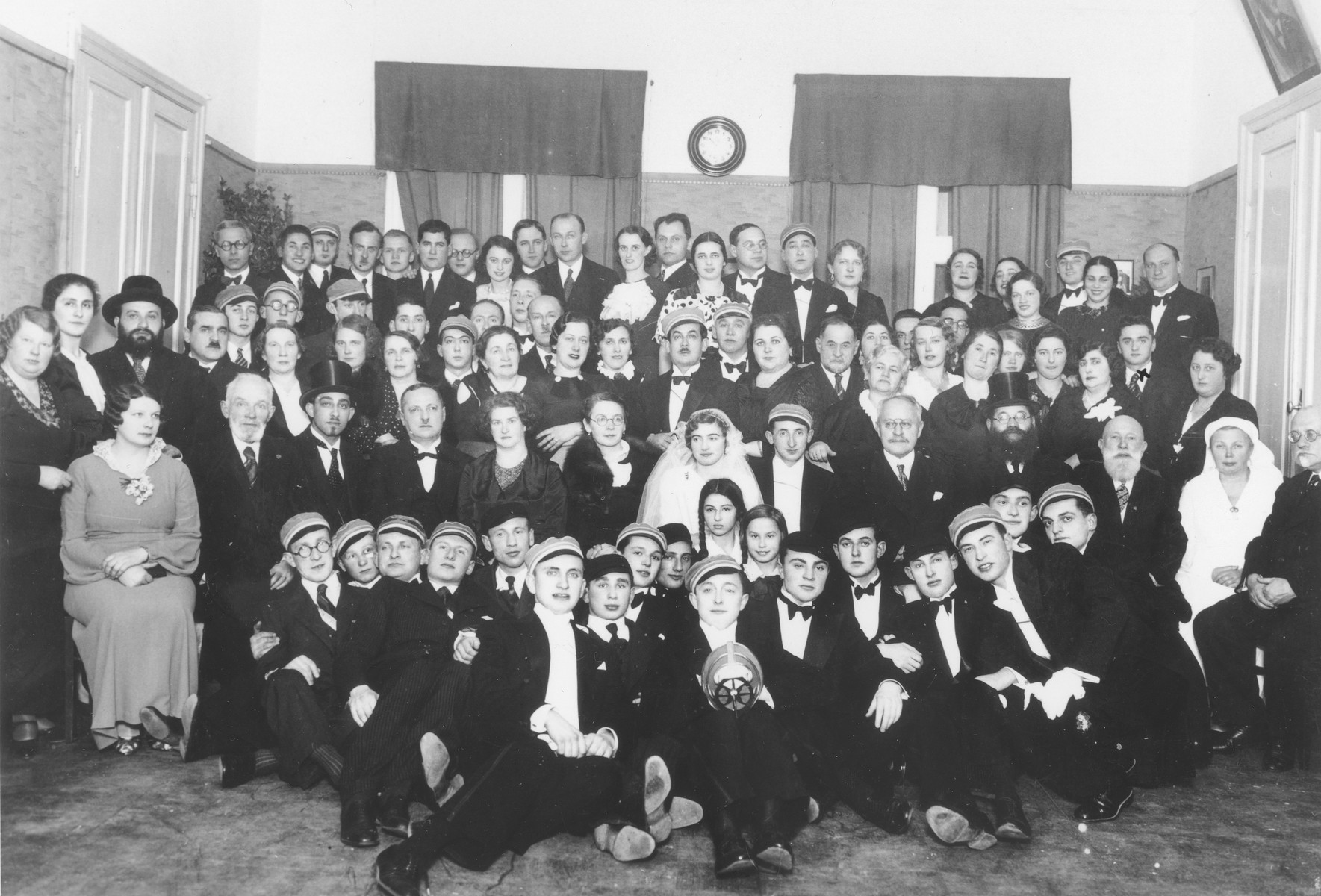 A Jewish bride and groom pose among a large group of family and friends at their wedding.

Many of the men are wearing the caps of a Jewish fencing club.