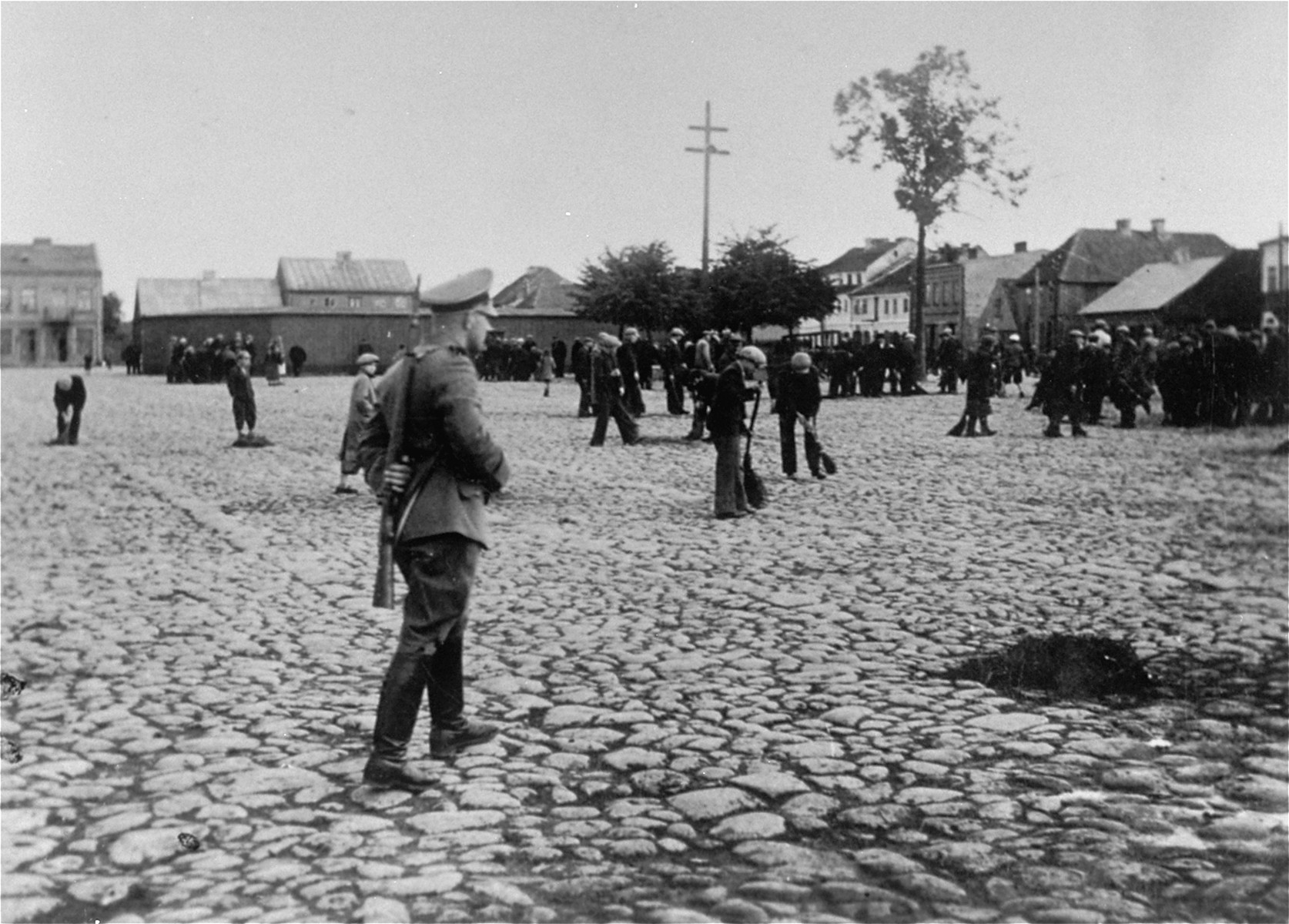 An SS member oversees a group of Jewish men at forced labor sweeping the pavement of the town square in Raciaz.