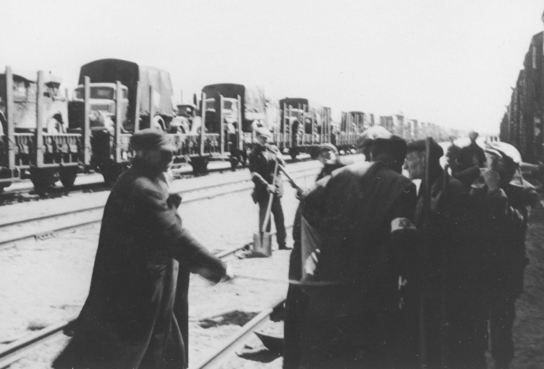 A group of Jewish men wearing armbands works at a railyard in Poland.

In the background is a long train carrying trucks.