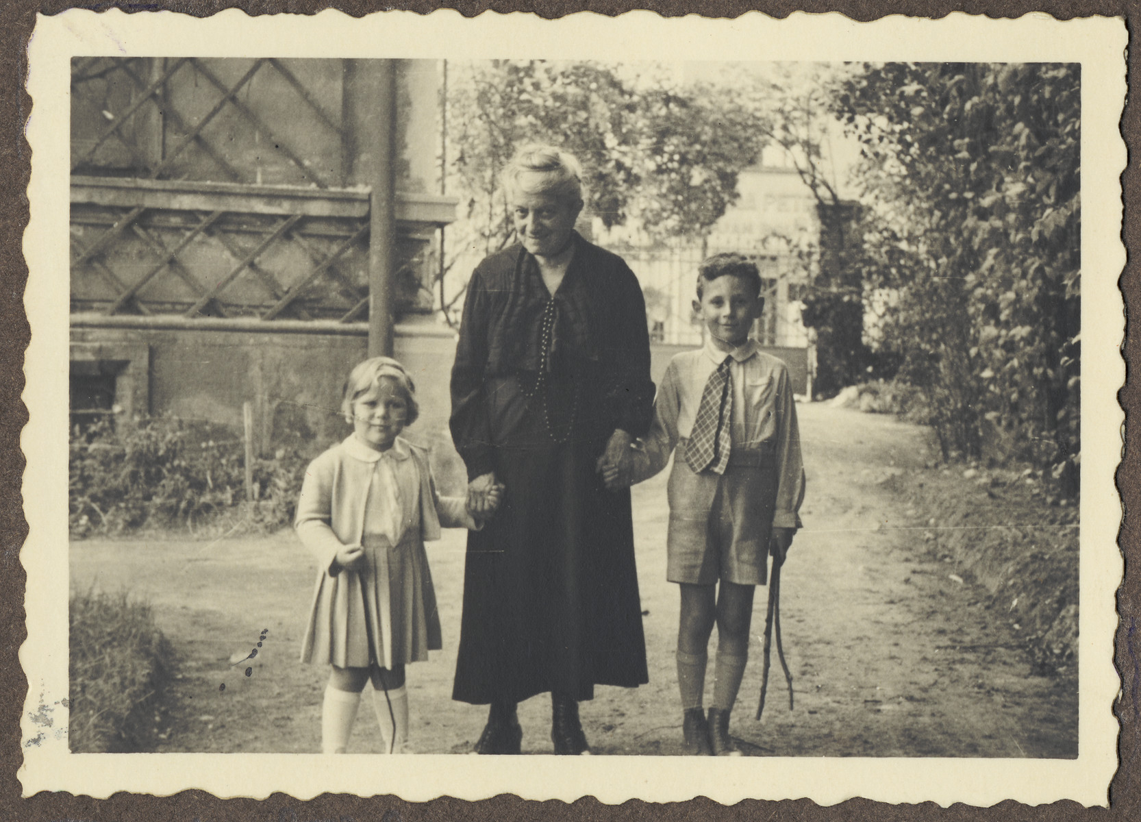 An elderly Czech Jewish woman walks through a garden with two young relatives.

Pictured on the right is Michal Kraus; on the left is his cousin Eva Schur.  The woman in the center is probably Eva's great grandmother.

Eva Schur, was the daughter of Franta Schur, the cousin of Michal Kraus.  Eva perished in Auschwitz with her mother and brother, while her father, Franta Schur survived the war in England.  The picture was taken in the garden of the house belonging to her grandmother, Jenny Schur.