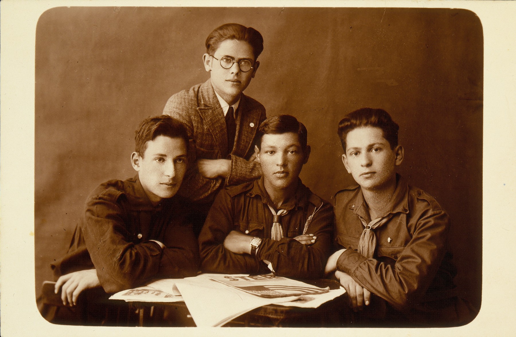 Four young members of the Betar youth movement gather around a table strewn with newspapers. 

Faivl Glombocki (sitting in center), on his right is Dov Wilenski, on his left Hanan Polaczek, and a friend  (standing) from Chiznistock, Vilna. Dov immigrated to Palestine.  Faivl came to Argentina in 1930 with his family and later immigrated to Israel.