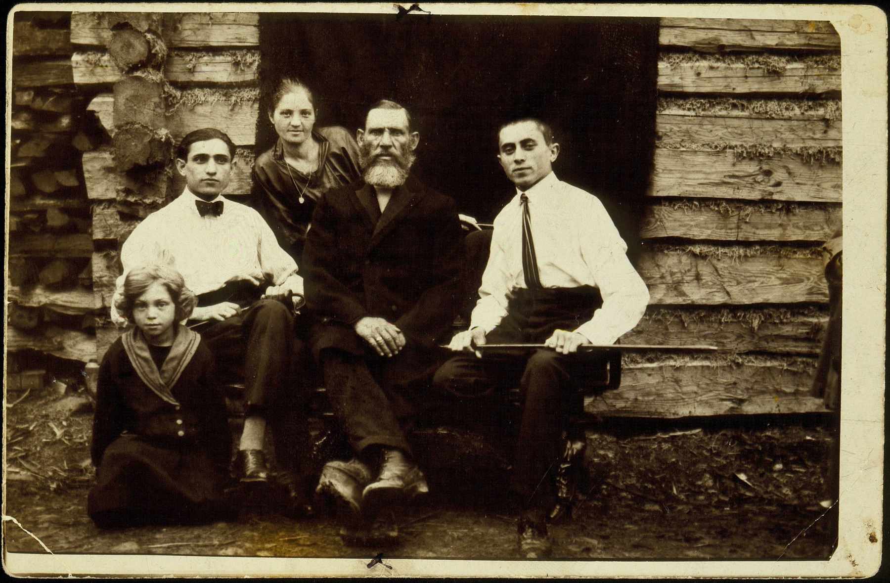 The Kremin family sits at the entrance to a log cabin. 

Yude-Mendl Kremin (center), with his son-in-law Hirshl Tatarski (right), daughter Rivka Tatarski, son Hayyim-Itchke, and granddaughter Hayya-Rochke Shmidt. 

Yude-Medl had a clubfoot as a result of a severe beating by Lida coachmen during a labor dispute.  Yude-Mendl died a natural death; all the others perished in the September 1941 massacre.