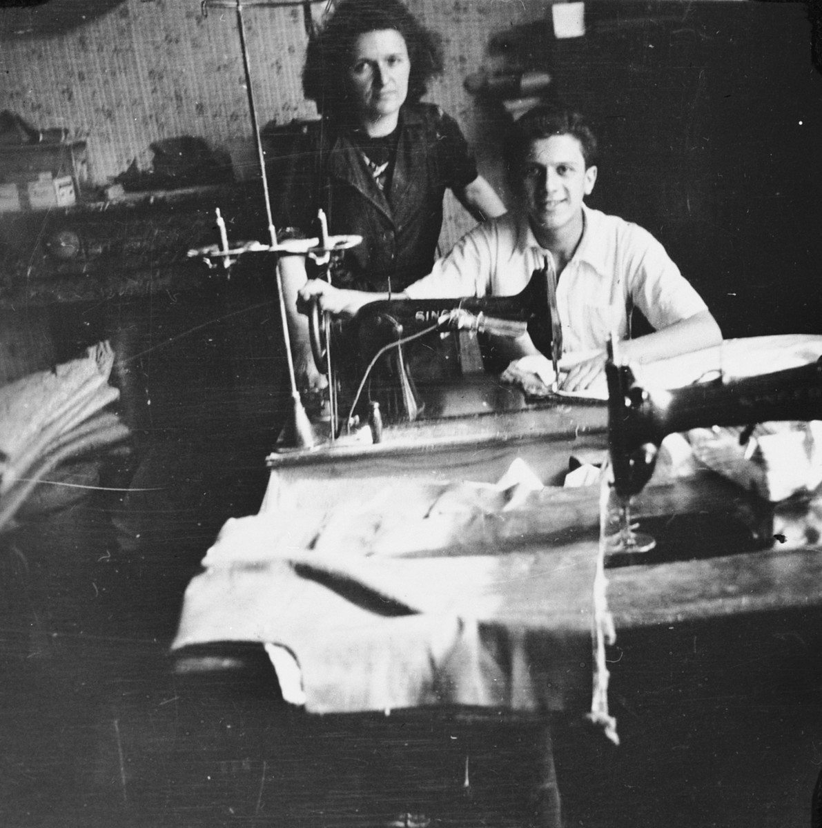 A Jewish woman who is living in hiding as a Christian in a factory in Brussels, poses with a young man who is seated at a sewing machine.

Pictured is Fani Mendelowicz.