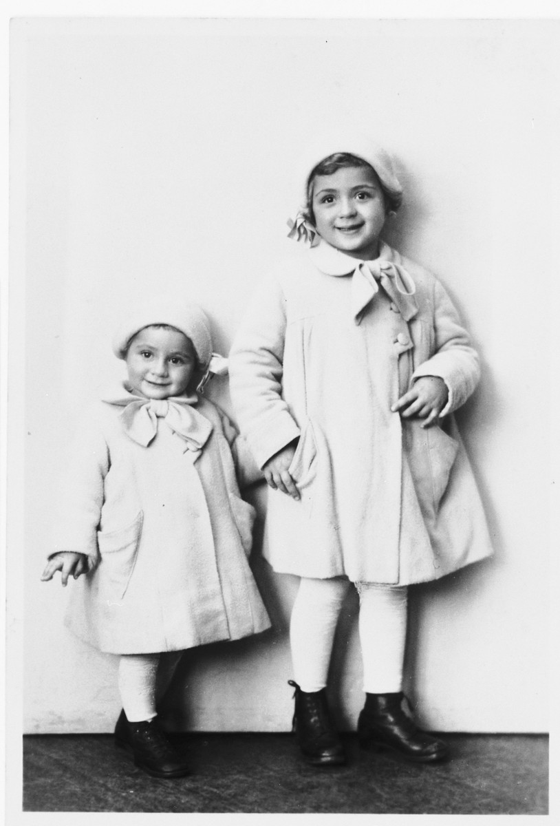 Studio portrait of two Jewish sisters dressed in matching coats and hats. 

Pictured are Miriam and Leah Spitzer.