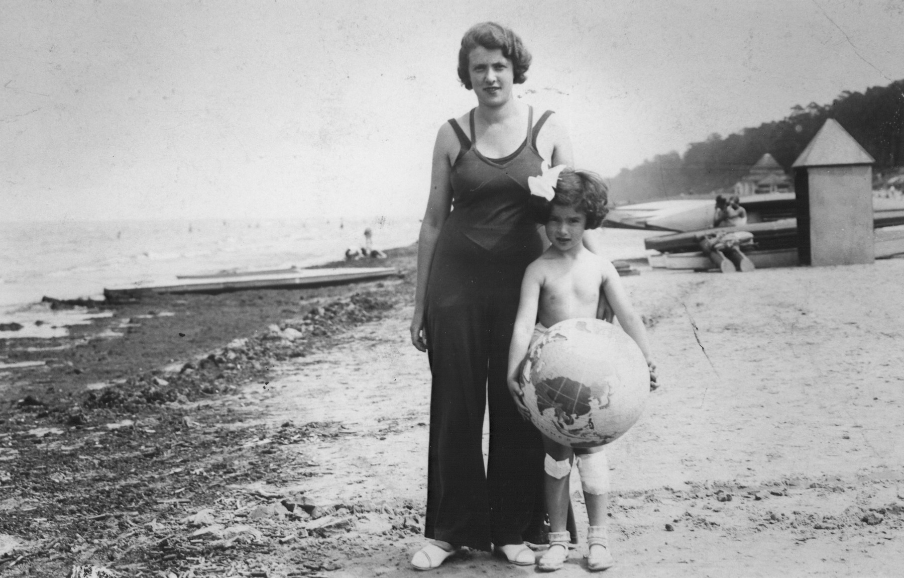 A young Jewish child holding a beach ball painted like a globe, poses with her mother on a beach, probably in Lithuania.

Pictured are Mira Jedwabnik and her mother, Lydia.