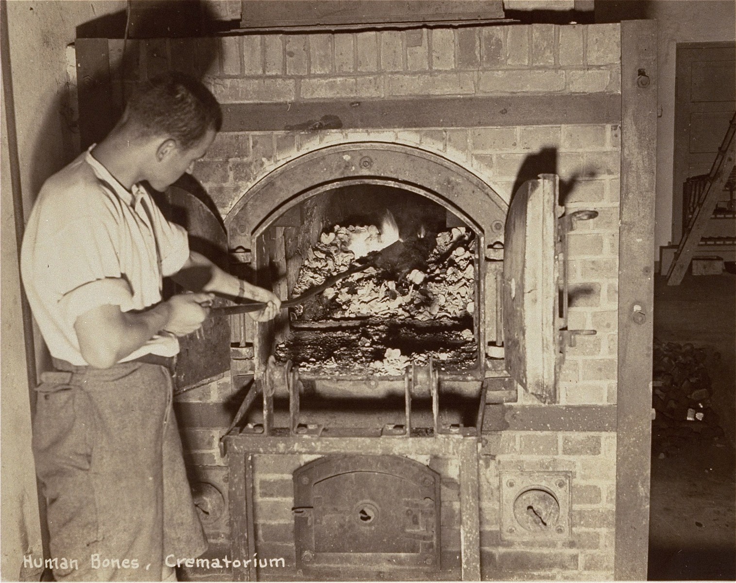 A survivor stokes smoldering human remains in a crematorium oven that is still lit.