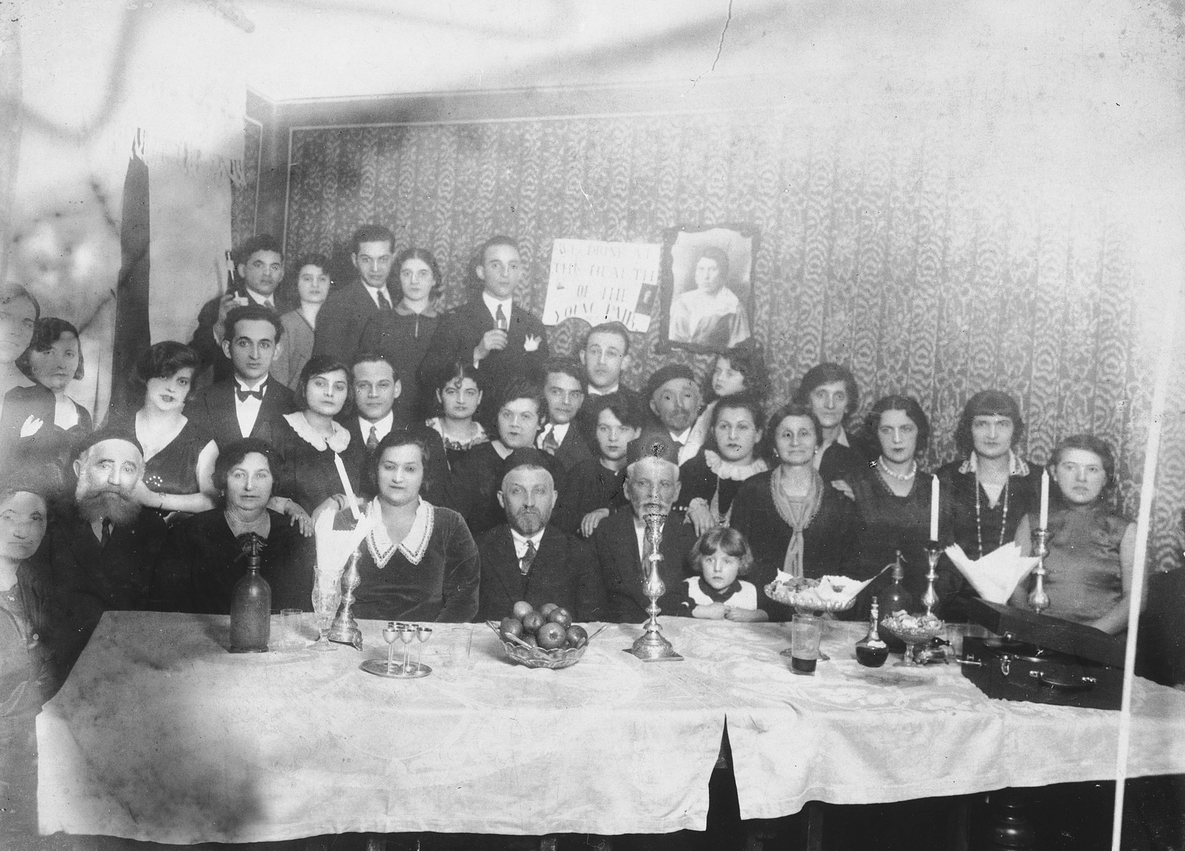 The Safirsztajn family gathers to celebrate the marriage of Mordechai Safirsztajn who had immigrated to the United States.

The sign hanging on wall behind them reads: "We drink to the health of the young couple."  Among those pictured is Regina Safirsztajn (far right), one of the four women who was hanged for her participation in the Auschwitz uprising. 

Pictured are last row, left to right: Ajzyk Safisrsztajn, unknown, [first name unknown] Gold, unknown, and [first name unknown] Gold.  Middle row: unknown, [first name unknown] Safirsztajn, wife of Wolf Safirsztajn, Wolf Safirsztajn, [first name unknown] Najmark, [first name unknown] Najmark, [first name unknown] Najmark, Chana Safirsztajn Ickowicz, Adek Eizel Safirsztajn, Abel Ickowicz, Szaja Ickowicz, Bronia Ickowicz, Tonia Safirsztajn, [first name unknown] Safirsztajn, "Mima", [first name unknown] Gold, [first name unknown] Gold, and Regina Safirsztajn.  Seated in first row, left to right: unknown, Icie Safirsztajn, Golda Safirsztajn, Karola Safirsztajn Najmark, Jakub Najmark, Josef Najmark Rozia Ickowicz.