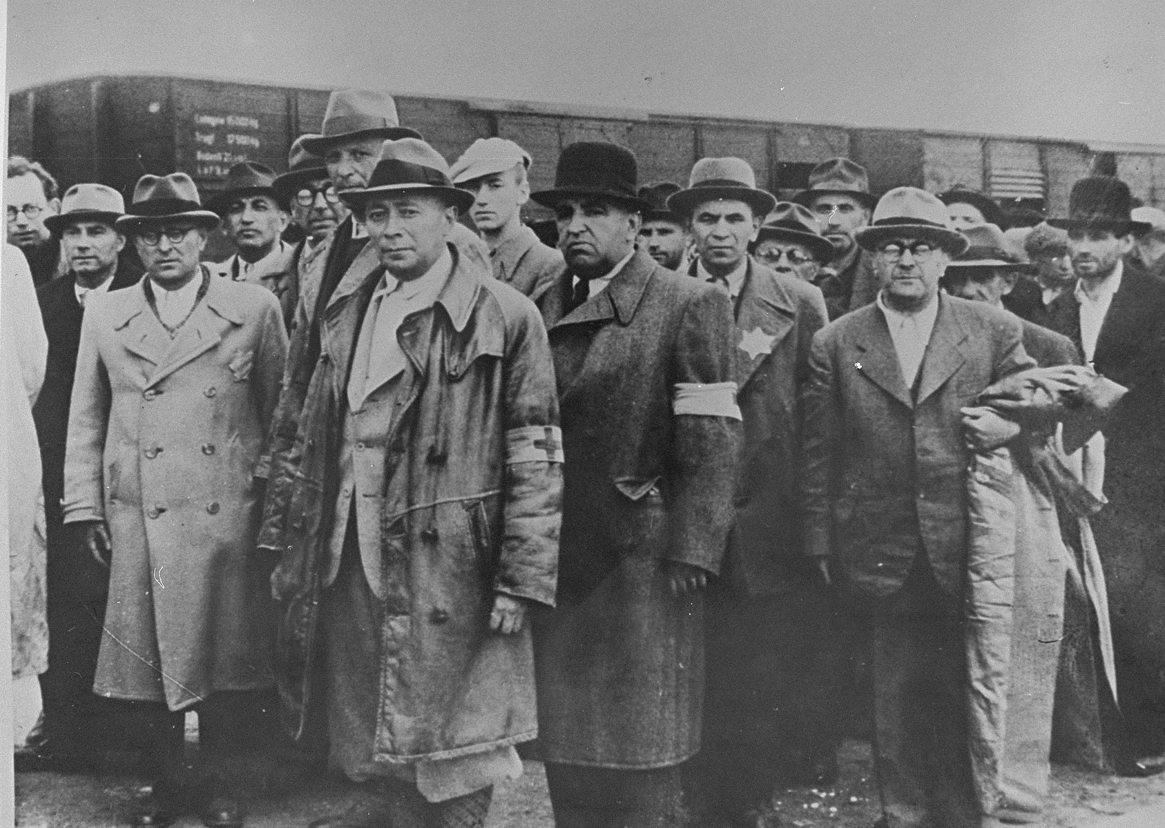 Jewish men from Subcarpathian Rus await selection on the ramp at Auschwitz-Birkenau. 

Among those pictured are: Weishouse, a pharmacist, (second from the left); Dr. Kellerman (next to Weishouse); Adolf Abraham (wearing glasses behind Kellerman);. Gyury Kellerman (in a white hat); Istvan Balasza, a pharmacist (front row, right).  All are from the town of Tecso.  Also pictured is Ludwig Granat (center) wearing an armband and black hat.

The only known to have survived is Istvan Balasza.