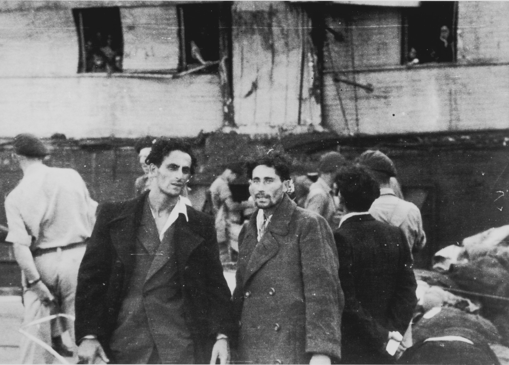 Two passengers from the illegal immigrant ship, Exodus 1947, stand on the dock in Haifa harbor in front of the battered ship.