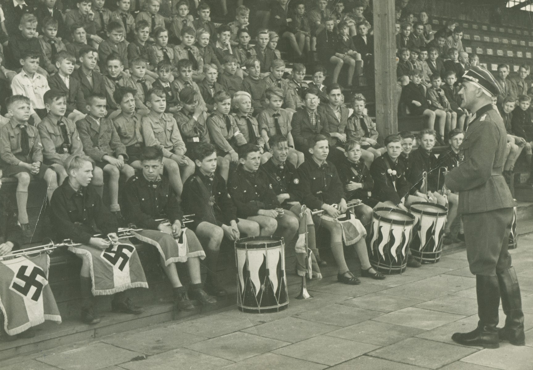 Young members of the Hitler Youth sit in a stadium behind a row of buglers and drummers.