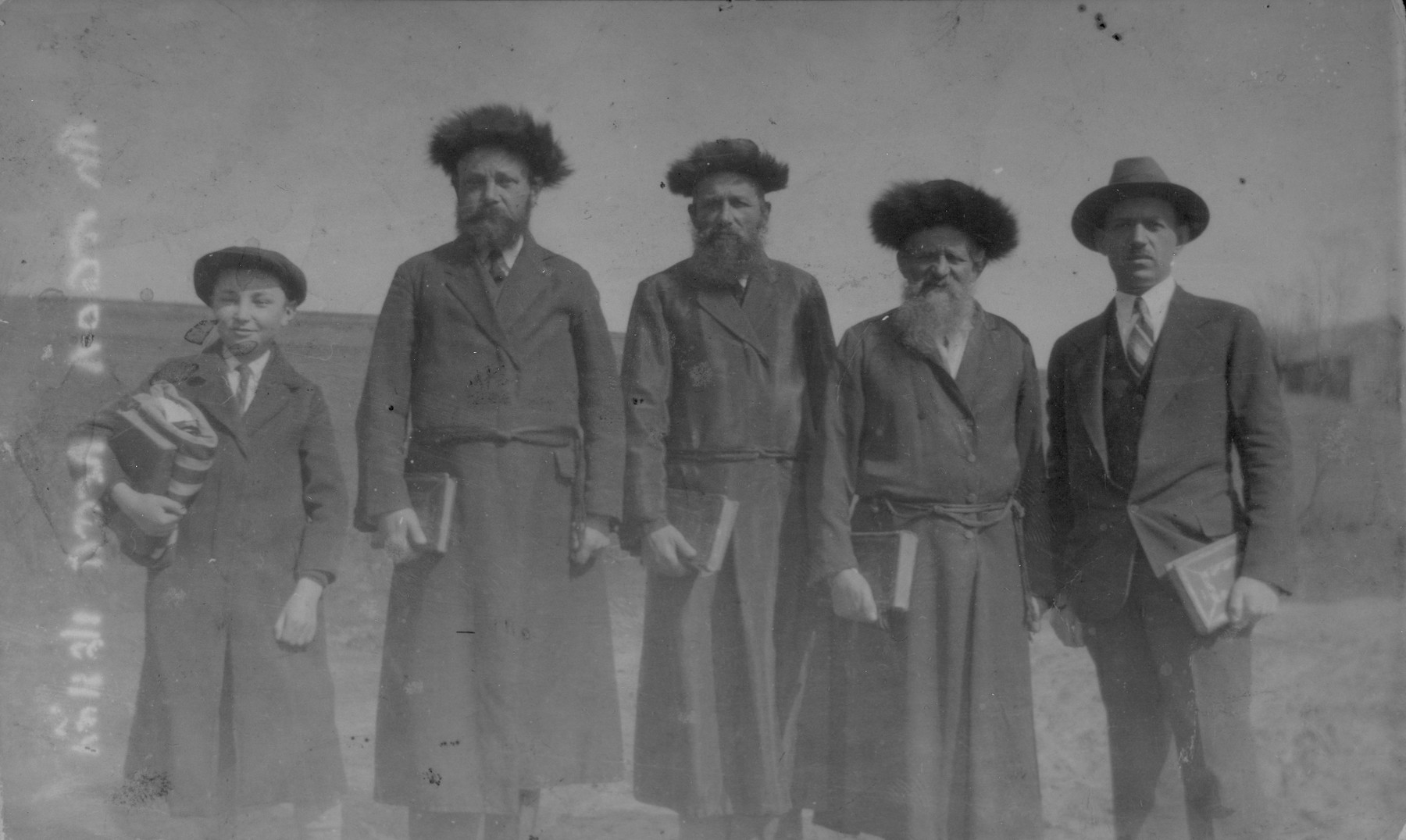 Group portrait of Hasidic Jews standing outside holding books in Raczyna, Poland.  

Pictured from left to right are: Chaim Yankel Gurfein, Lejzer Gurfein, Moshe Yosl Dudlsack, Itzhak Dudlsack, and an unidentified American visitor.