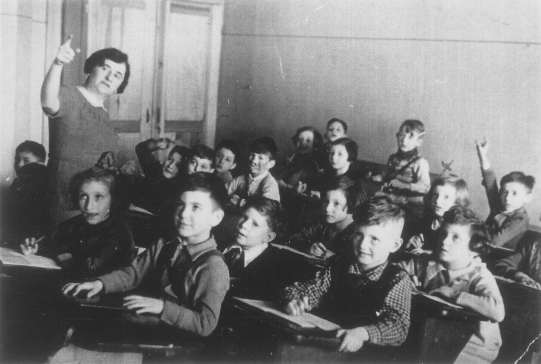 Students sit at their desks in a first grade classroom in Berlin.

Hannelore Mansbacher is sitting on the right side in front of the child who is holding up his hand. The teacher, Hilde Goldschmitt, later emigrated to Israel.