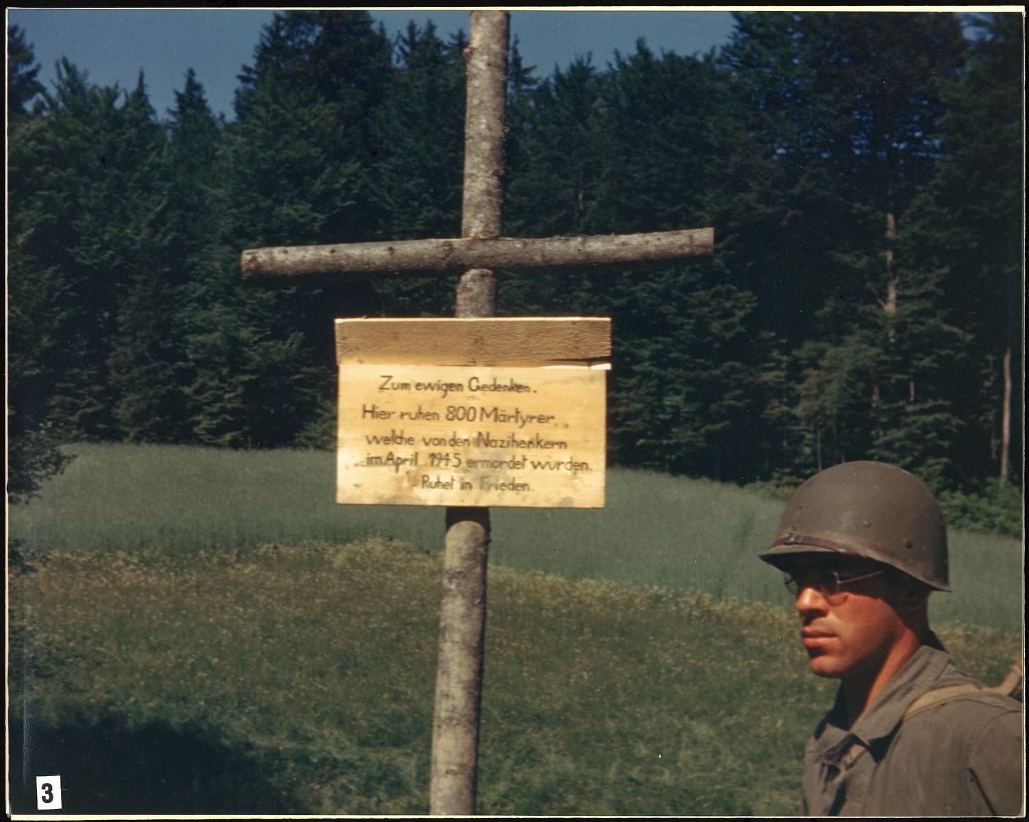 An American soldier stands next to a sign erected by the U.S. Army to mark the site of the Nammering atrocity.  It reads: "In eternal memory.  Here lie 800 martyrs who were murdered by Nazi executioners in April 1945.  Rest in peace."