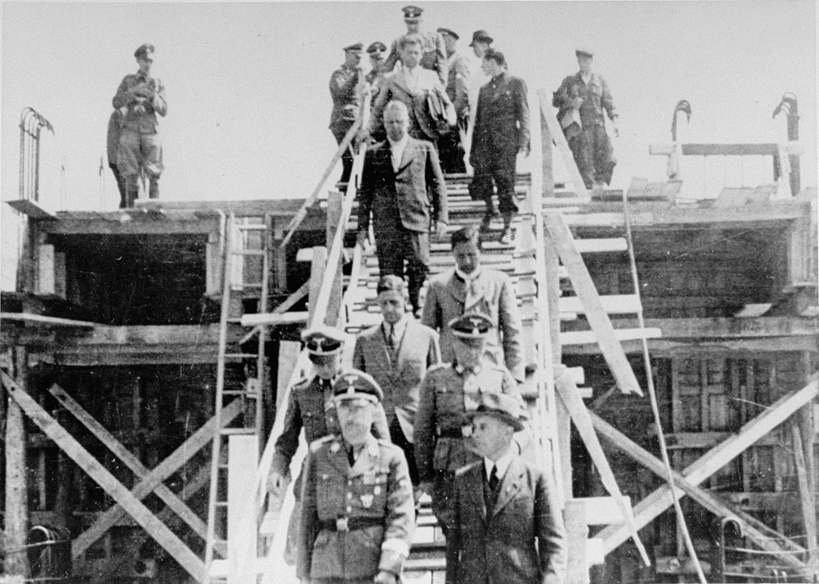 Reichsfuehrer SS Heinrich Himmler (bottom left) descends a wooden staircase during a tour of the Monowitz-Buna building site in the company of Max Faust (bottom right). 

Faust, who was an IG Farben engineer, was the head of building operations at Monowitz-Buna.