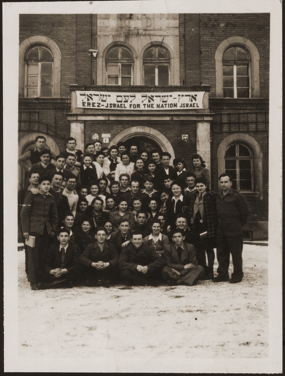 A group of Jewish DPs pose in front of a building in the Deggendorf DP camp. 

The banner reads in Hebrew and English, Erez Israel for the nation Israel (Land of Israel for the People of Israel).  The group, instructed by members of the Jewish Brigade,  was a small, centrist, slightly left-of-center Zionist movement called Noam (literally "pleasantness").  Shortly after the photo was taken members of the group formed an agricultural kibbutz to prepare for their aliyah to Israel.  Pictured in the photo are Hadasa Cudzynowska, Henik and Sala Weingarten.