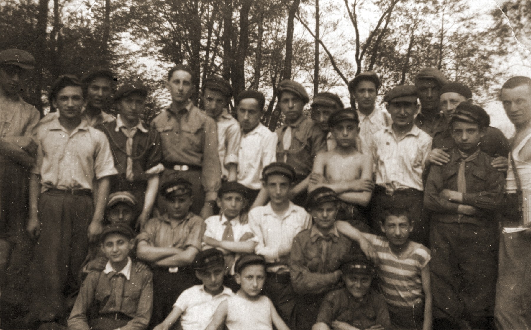 Group portrait of members of the Hashomer Hadati Zionist youth organization on an outing in the woods.

Pictured (top row, from left to right): unknown; Blumenfeld; unknown; Szmuel (Samuel) Gottlieb; Motek Rechnic; Motek Zajac; unknown; Hesiek Rechnic; Motek Ostrowiecki; Ilek Stawski;  Majer Nowomiejski; Szolem Blumenfeld; Mojszele Gruszka; Nordon, and Menachem Bukowski; (second row, left to right) are: Chune Kenner; Icek Rendel; Josek Lenczner; Rueven Lefkowicz; Motek Rendel, and Moniek Rozen (the donor, in striped shirt, aged 13 years);  (front row, from left to right) are: Hercko Lenczner; Jakub Bajtner; Moniek Szeps, and Motek Kenner.