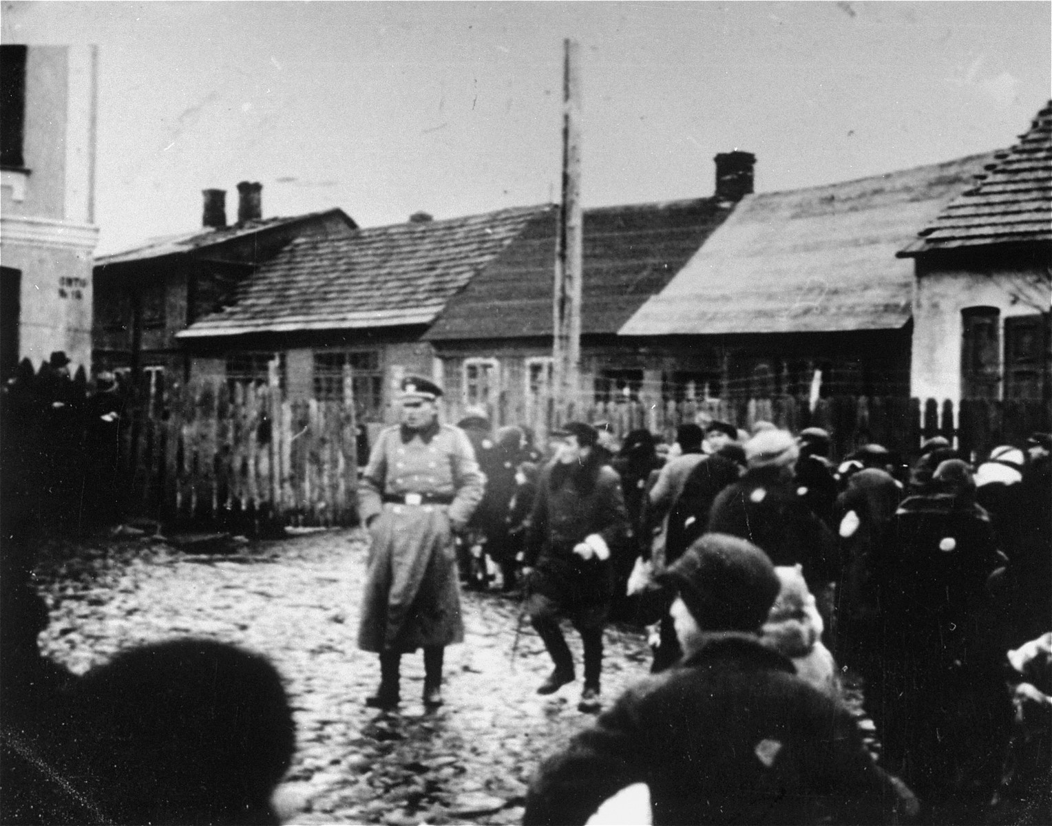 A German police officer stands in a fenced-in yard among a group of Jews, who have been rounded-up in the Ciechanow ghetto.