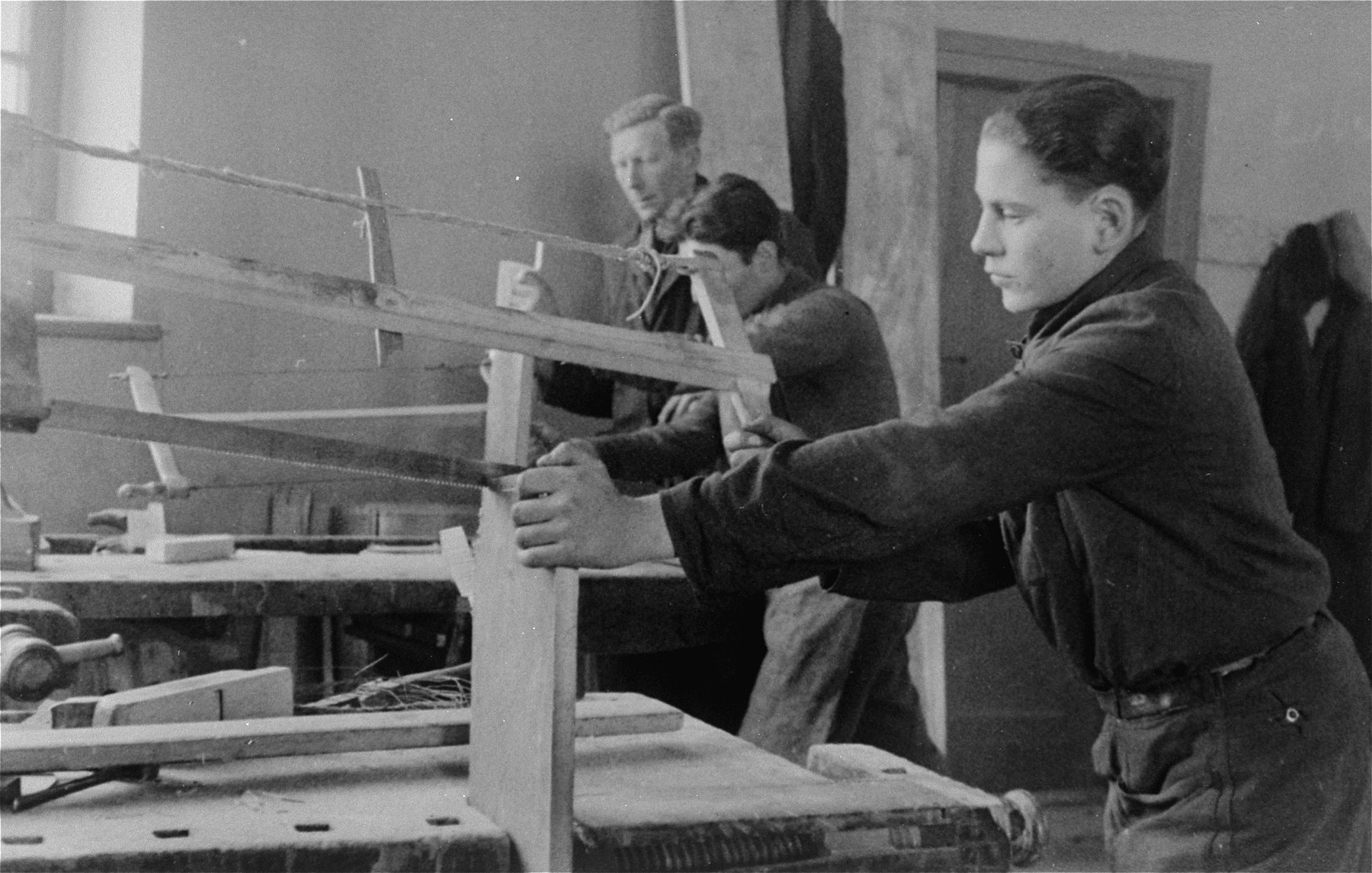 Jewish youth saw pieces of lumber in a carpentry workshop in the Bochnia ghetto.

The original German caption reads: "City workshops in Bochnia.  Jewish labor strength made useful." 

Pictured in the foreground is Romek Abraham Marber.
The boy with his face covered is Matzner  and the man in back is the workshop manager, Ginsberg or Greenberg.  He was an architect from Krakow.