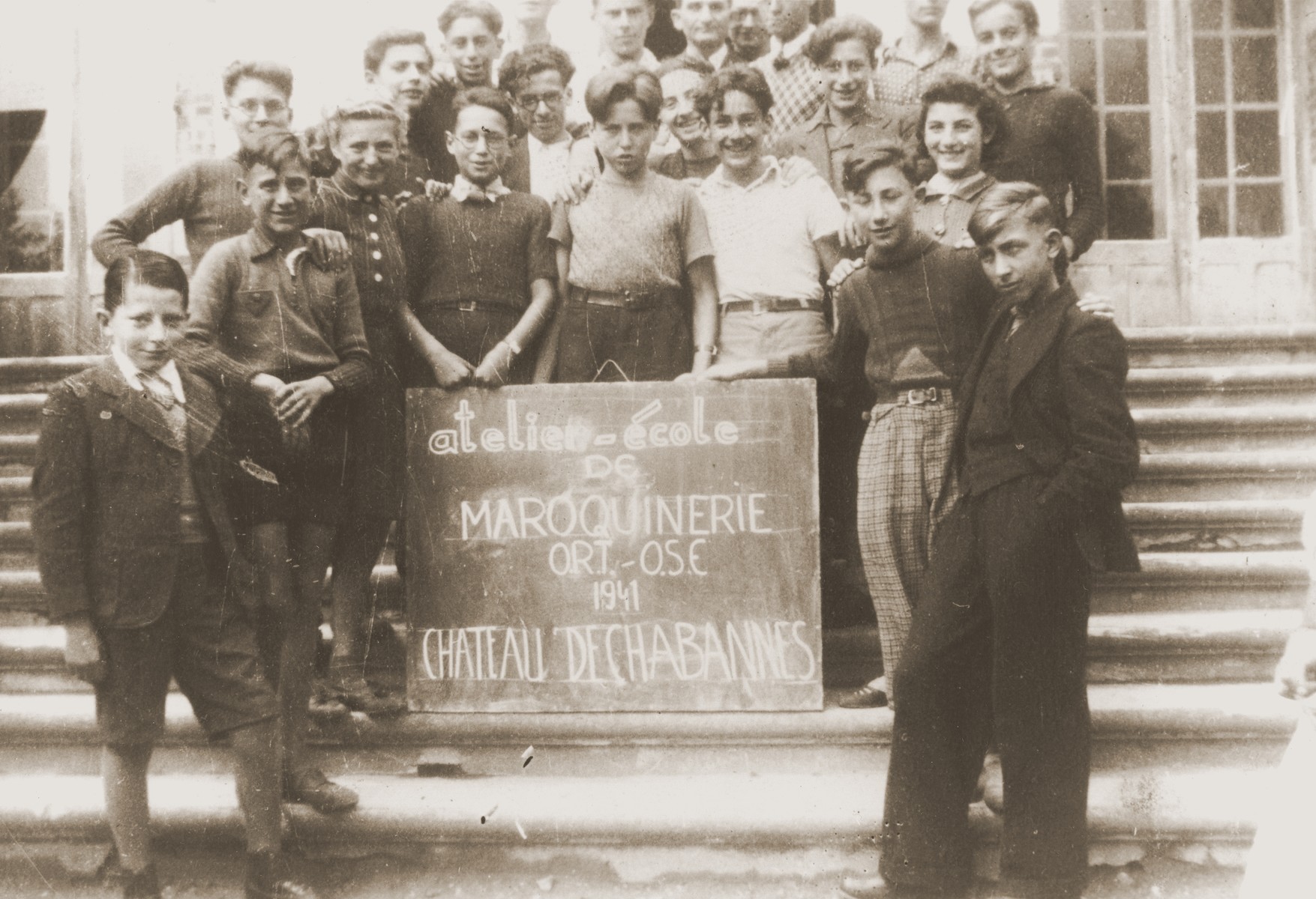 Jewish refugee youth who are students in an ORT leather working class at the Château de Chabannes children's home, pose on the front staircase of the home.  Château de Chabannes was sponsored by the OSE (Oeuvre de secours aux Enfants).  

Heinz Stephan Lewy is pictured in the front row, center.  Egon Halbright (born Halbreich) is located in the second row, wearing glasses and a white shirt--identified by his daughter, Rita Halbright