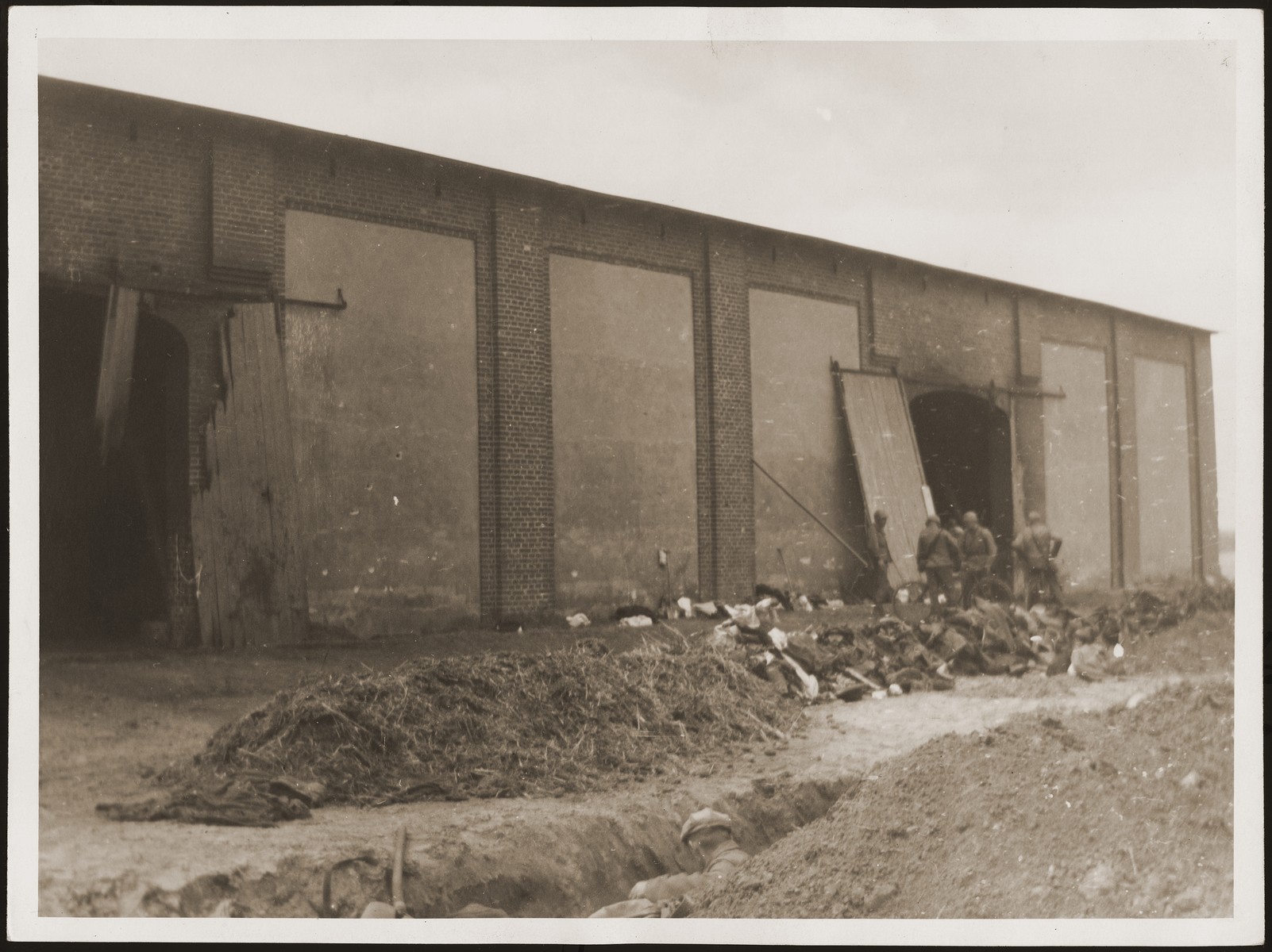 American soldiers stand in front of the barn in which concentration camp prisoners were burned alive by the SS.  German civilians exhume a mass grave in the foreground.