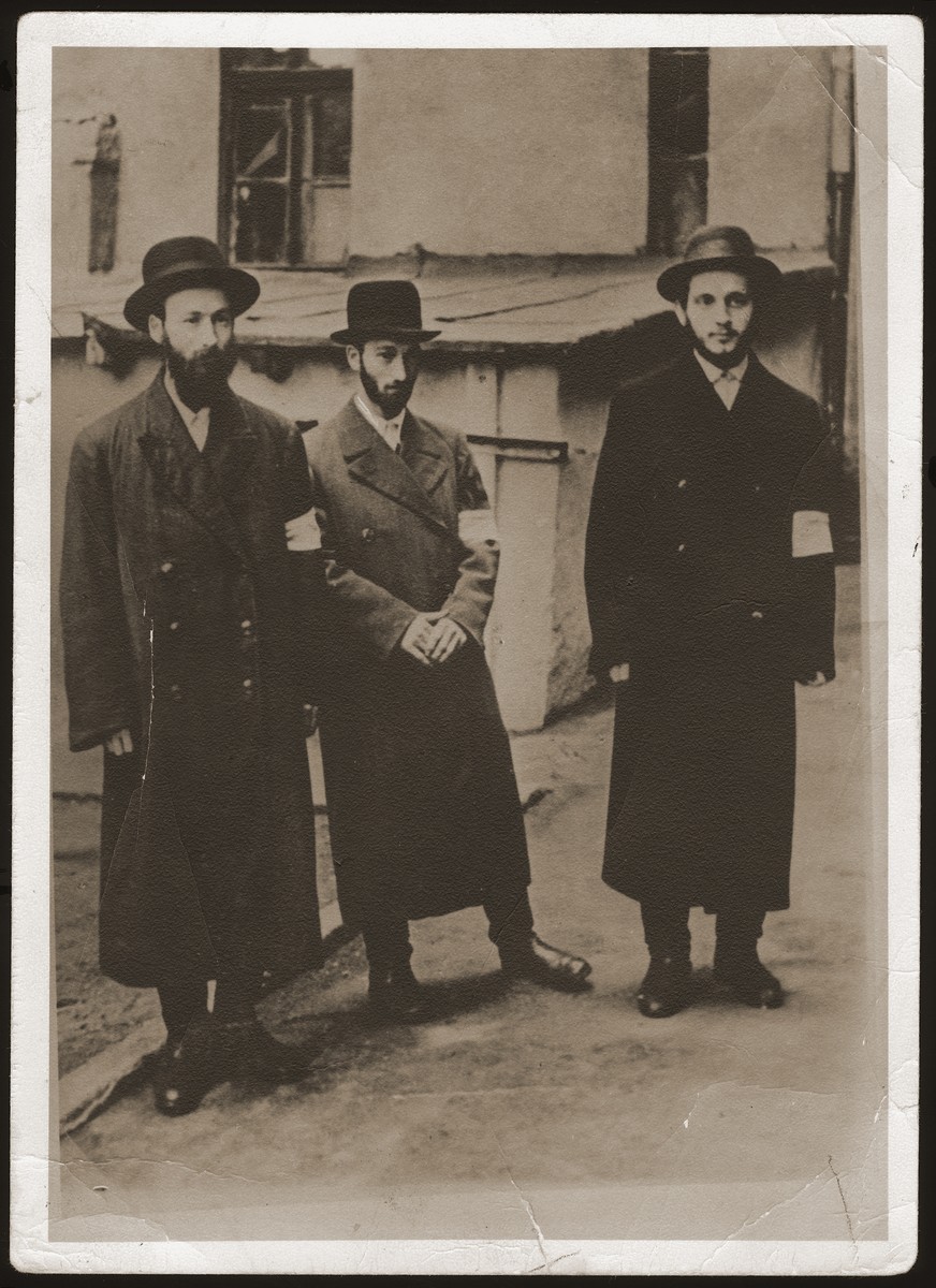 Three religious Jewish men wearing armbands pose on a street in the Bedzin ghetto.

Pictured from left to right are: Szlojme Werdygier, Lejbl Werdygier and Josl Lask.  The Werdygier brothers perished in Auschwitz.