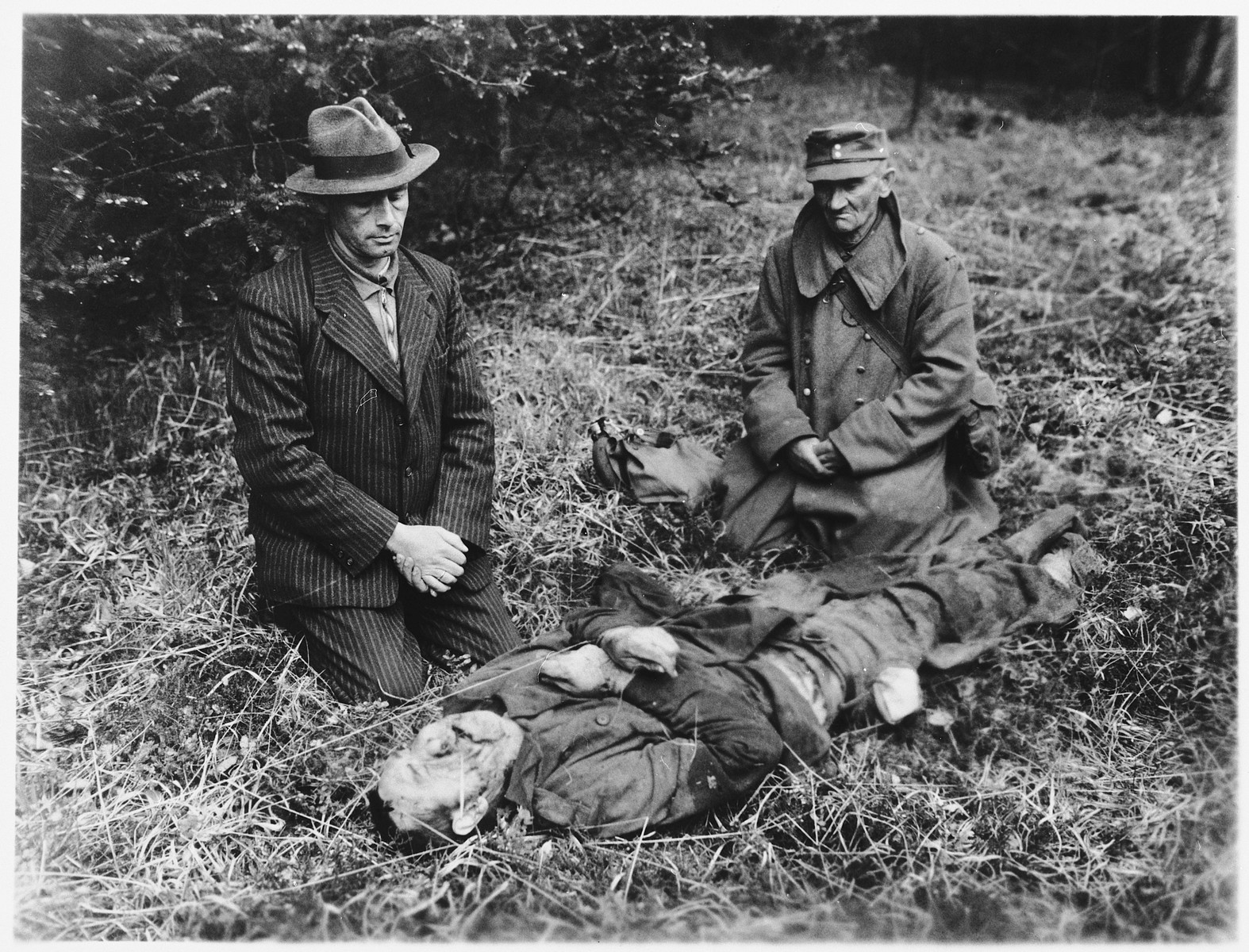 A Nazi party member from Hals and a German prison guard kneel beside the body of a Russian slave laborer who was killed by SS troops near Tiefenbach, near Passau. 

They were forced to view the atrocity by U.S. Third Army troops.  The victim was one of 100 Russians interned in a barn in Oberjacking, a small village near Tiefenbach.