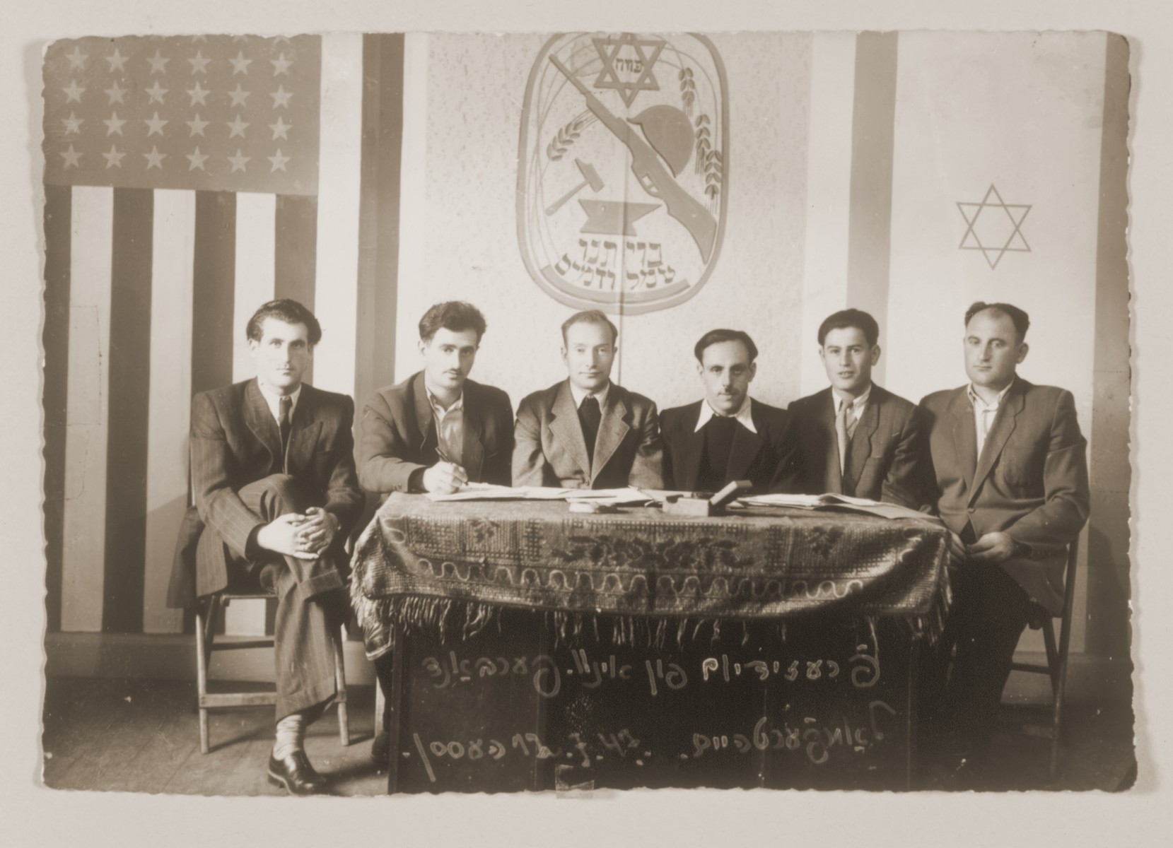 Meeting of the six-member presidium of an association in the Lampertheim displaced persons camp.  

The shield above the table bears the motto, "Our covenant, toil and blood."
