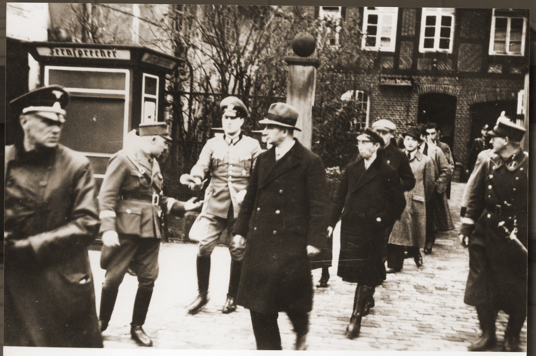 A small group of Jewish men, who have been rounded-up for arrest in the days after Kristallnacht, file out of the police station in Stadthagen, escorted by German police and SA members.