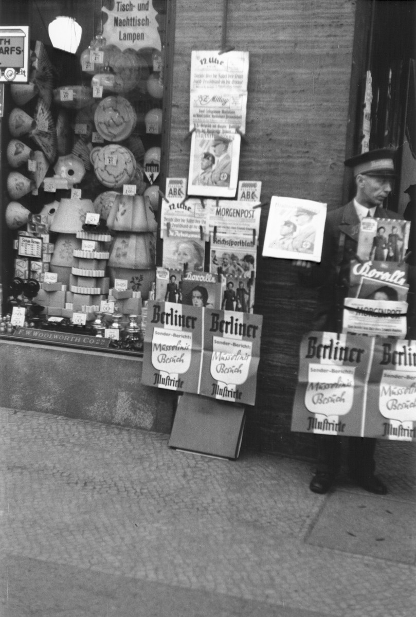 A newspaper vendor stands next to a display of German newspapers and magazines on a commercial street in Berlin.  

The advertisement for the Berliner Illustrirte features coverage of Mussolini's official visit to Berlin in September 1937.
