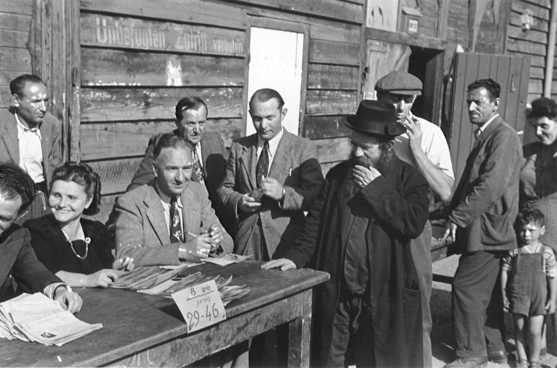 Jewish DPs line up in front of a wooden barracks to register with IRO (International Relief Organization) officials.  

The Hebrew sign on the table reads, "Table B, Barracks 29-46."