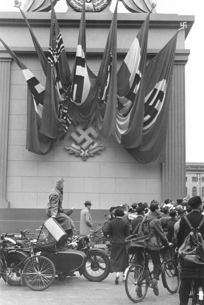 German spectators at a Nazi rally stand alongside a monument decorated with Nazi flags and a swastika emblem in Berlin.