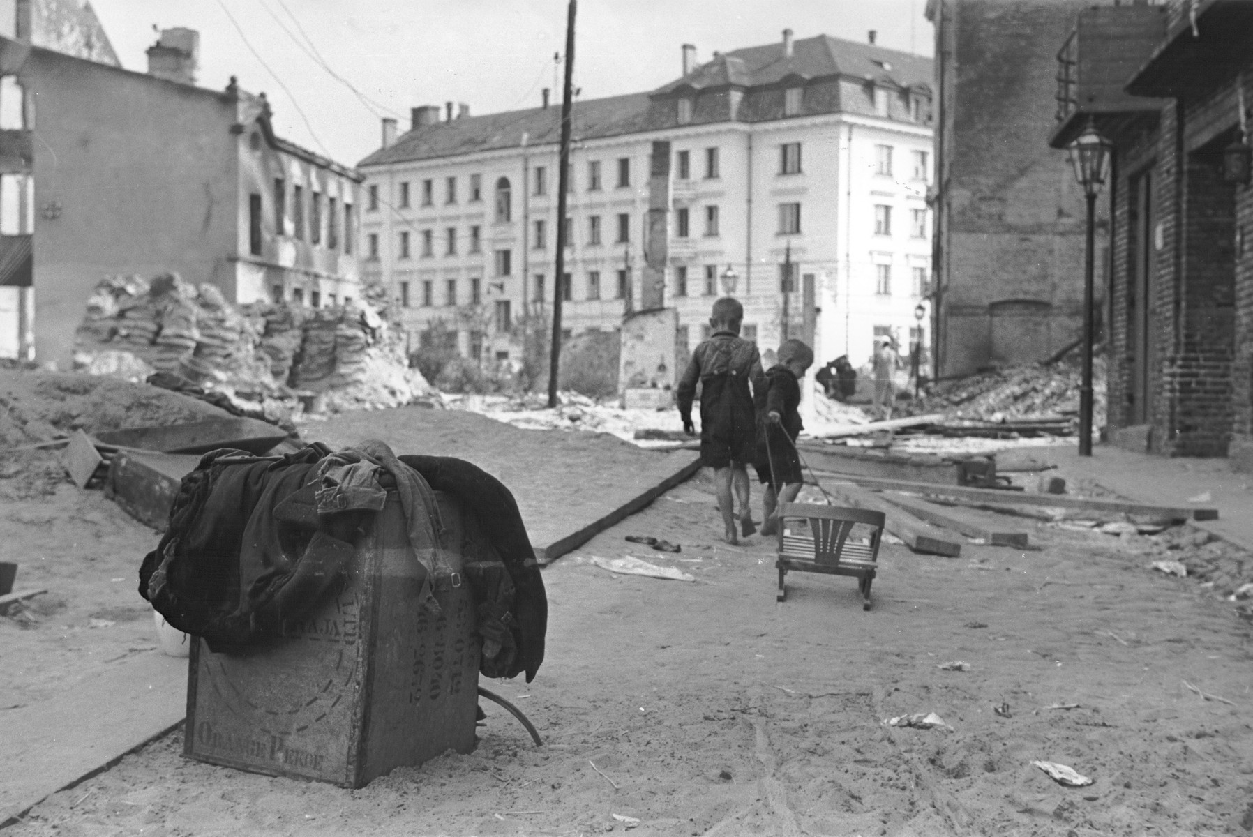 Two young Polish children pull a toy chair along a street in Warsaw that is strewn with rubble as a result of German air raids.

In the foreground is an abandoned crate stamped with the words "Orange Pekoe."