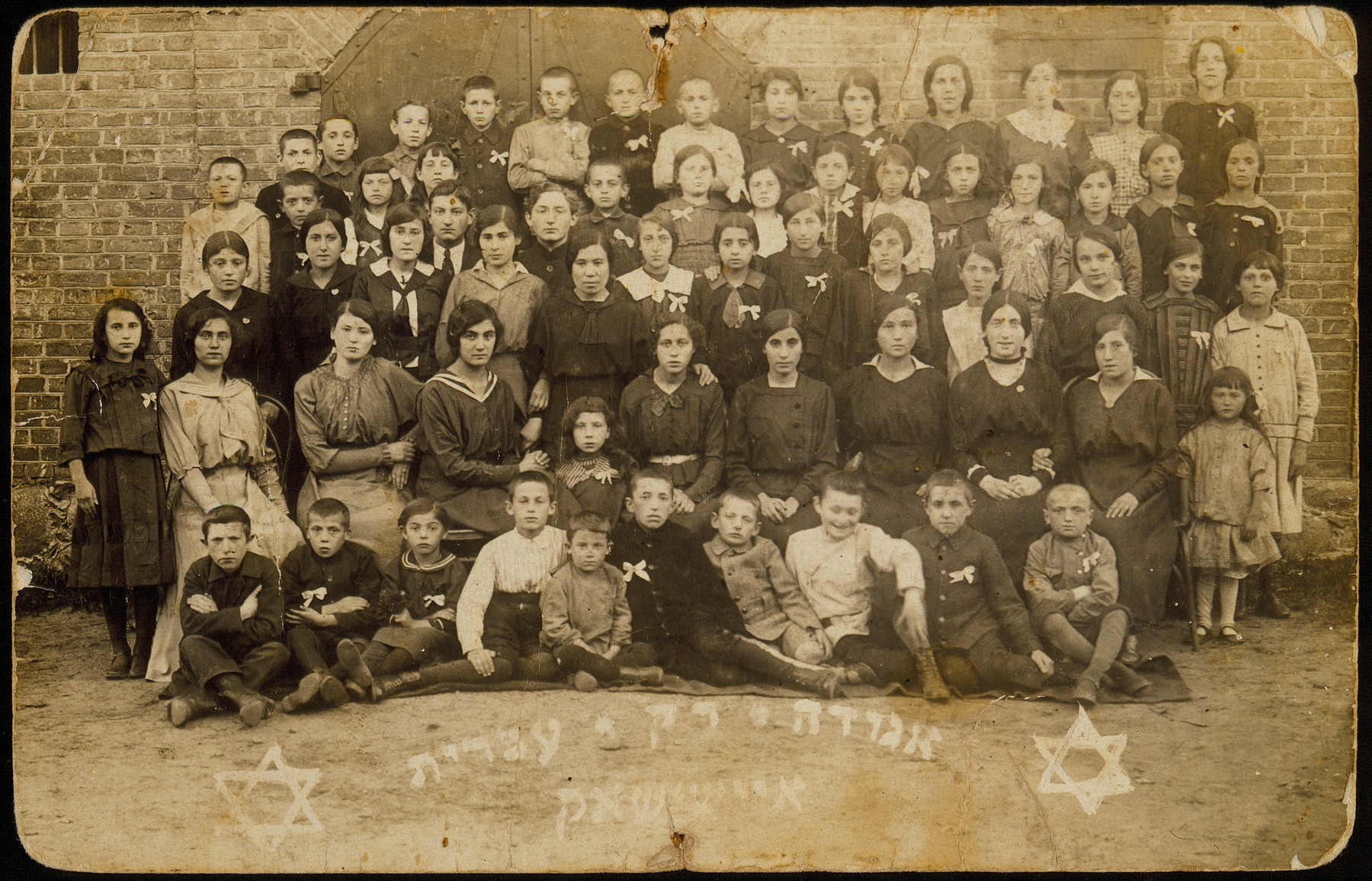Members of the Rak Ivrit (Only Hebrew) society founded by Leah Wilansky. 

The white bow on their chests indicates that Hebrew, not Yiddish, is the member's daily language.  In the third row, fifth from left, is Sarah Rubinstein, the Hebrew instructor, who was the first female teacher to teach boys as well as girls.  

Shlomo Kiuchefski is sitting on the ground, fifth from right.  The little girl in the second row, far right, is Esther Katz.  Fourth row, standing to the right of Sarah Rubinstein, is Shaul Kaleko and his brother Simha; the boy directly behind Sarah is Moshe Sonenson.  Behind him in the last last row is his brother Shalom, wearing a black outfit.  A significant number of the people in the photo immigrated to Palestine or other various countries.