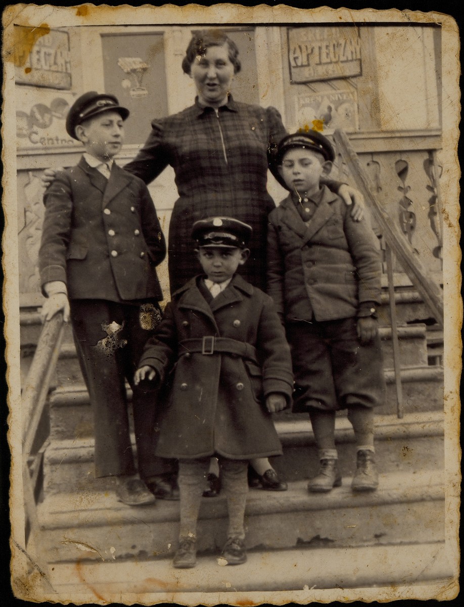 Alte Katz stands on the steps of her drugstore flanked by three young relatives. 

Alte Katz is in the center. To her right is her youngest son Avigdor.  To her left is her oldest grandson Yitzhak Sonenson.  In front of her is one of her great nephews Shaul Replianski, the son of Esther (nee Dwilanski) and Mordekhai Replianski. Shaul was the grandson of Shmuel Dwilanski. 

Alte Katz, her son Avigdor, and Shaul were murdered by the Germans during the September 1941 mass shooting action in Eisiskes.  Yitzhak survived the Holocaust in hiding.