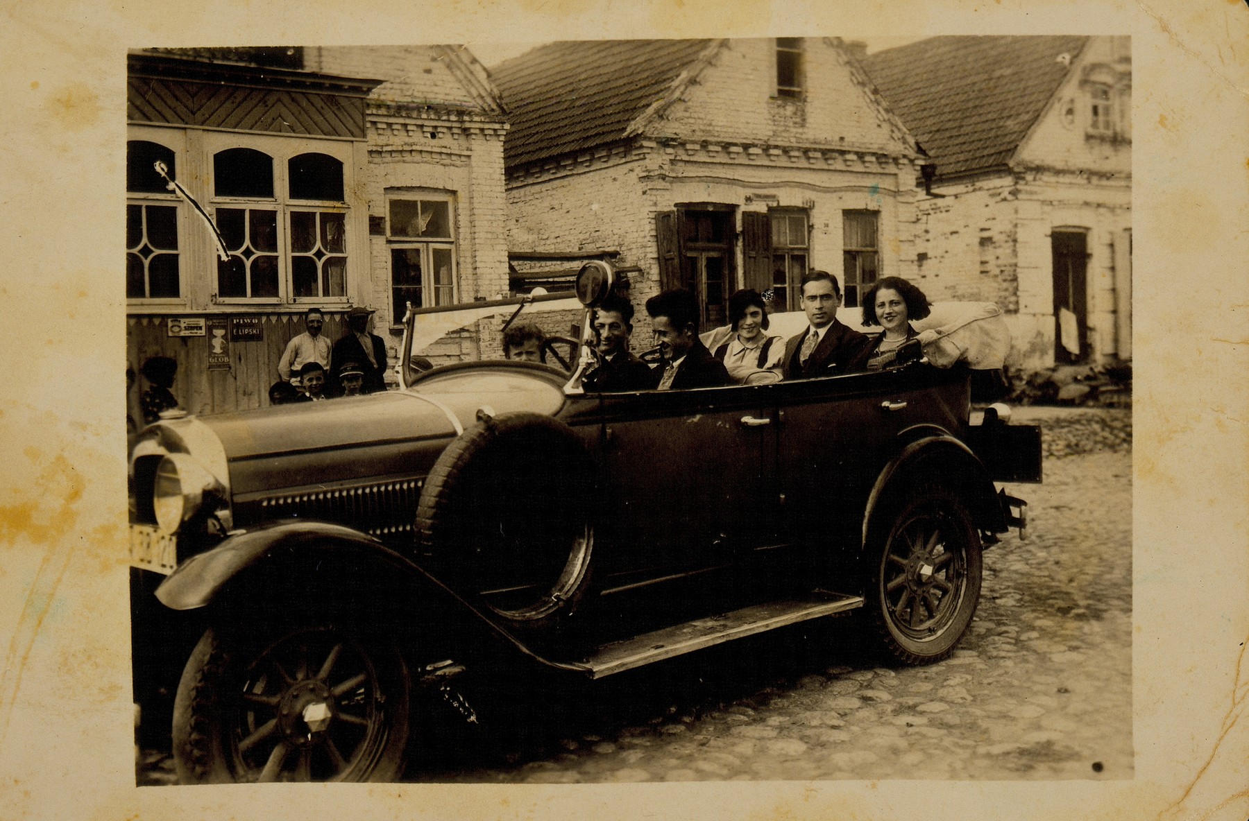 A visitor to Eisiskes takes his friends for a car ride in the early 1930s. 

Seated next to the driver is Leibke Sonenson and his nephew Benyamin.  In the back seat (left to right) are Matle Sonenson Shereshefski, Shaul Dubrowicz and the driver's friend.  The little boy without the hat on the extreme left is Leibke Kaganowicz (Leon Kahn).  The houses in the background belong to the Kaganowicz and Blacharowicz families.  Matle survived the war in Siberia; all the others, except for Leon, perished during the Holocaust.