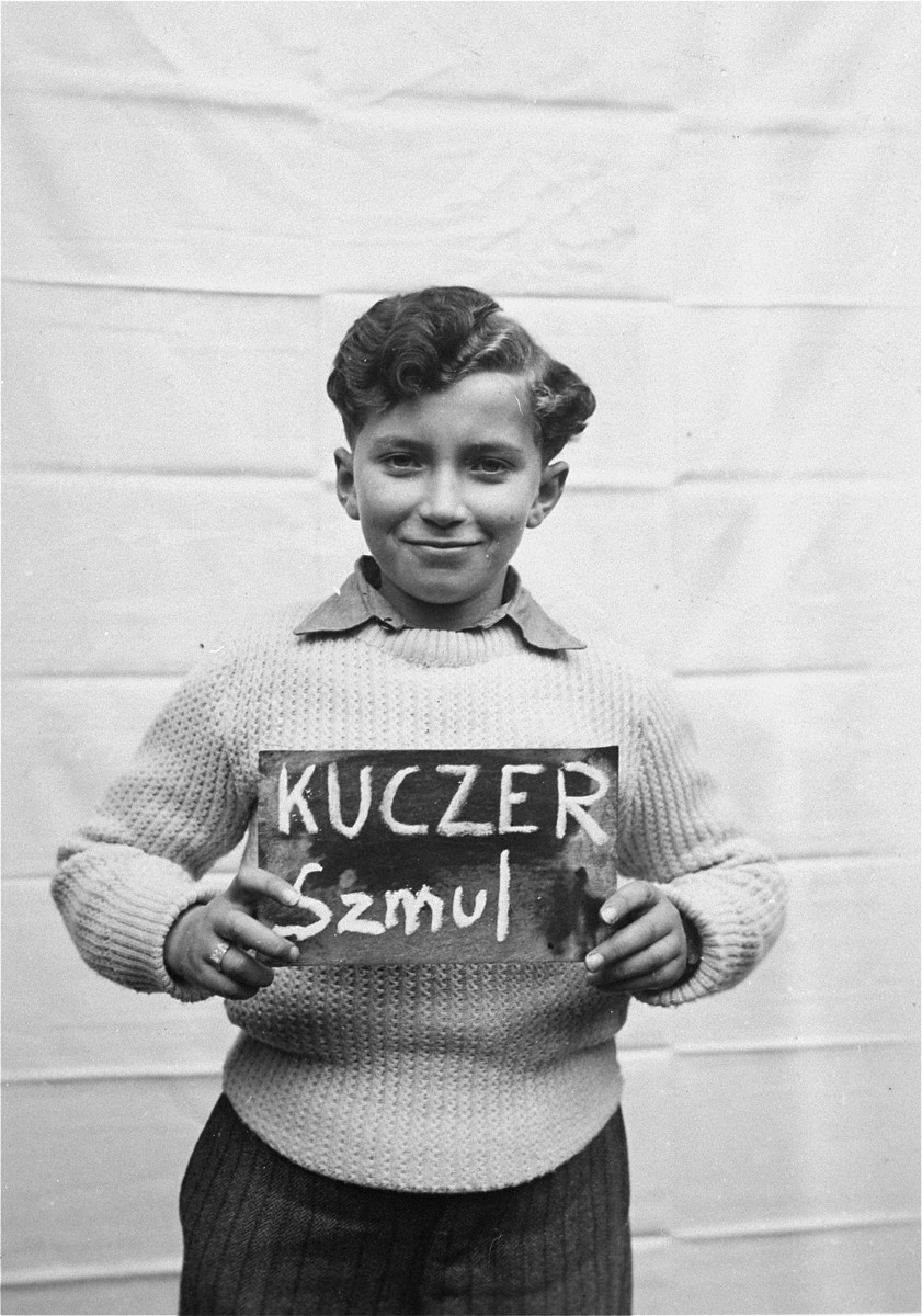 Szmul Kuczer holds a name card intended to help any of his surviving family members locate him at the Kloster Indersdorf DP camp.  

This photograph was published in newspapers to facilitate reuniting the family.